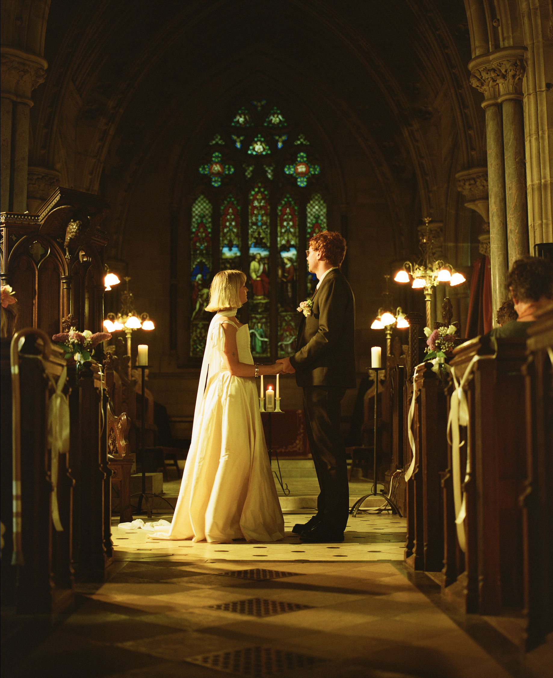 A couple getting married in a church, holding hands and facing each other with a stained glass window behind them.