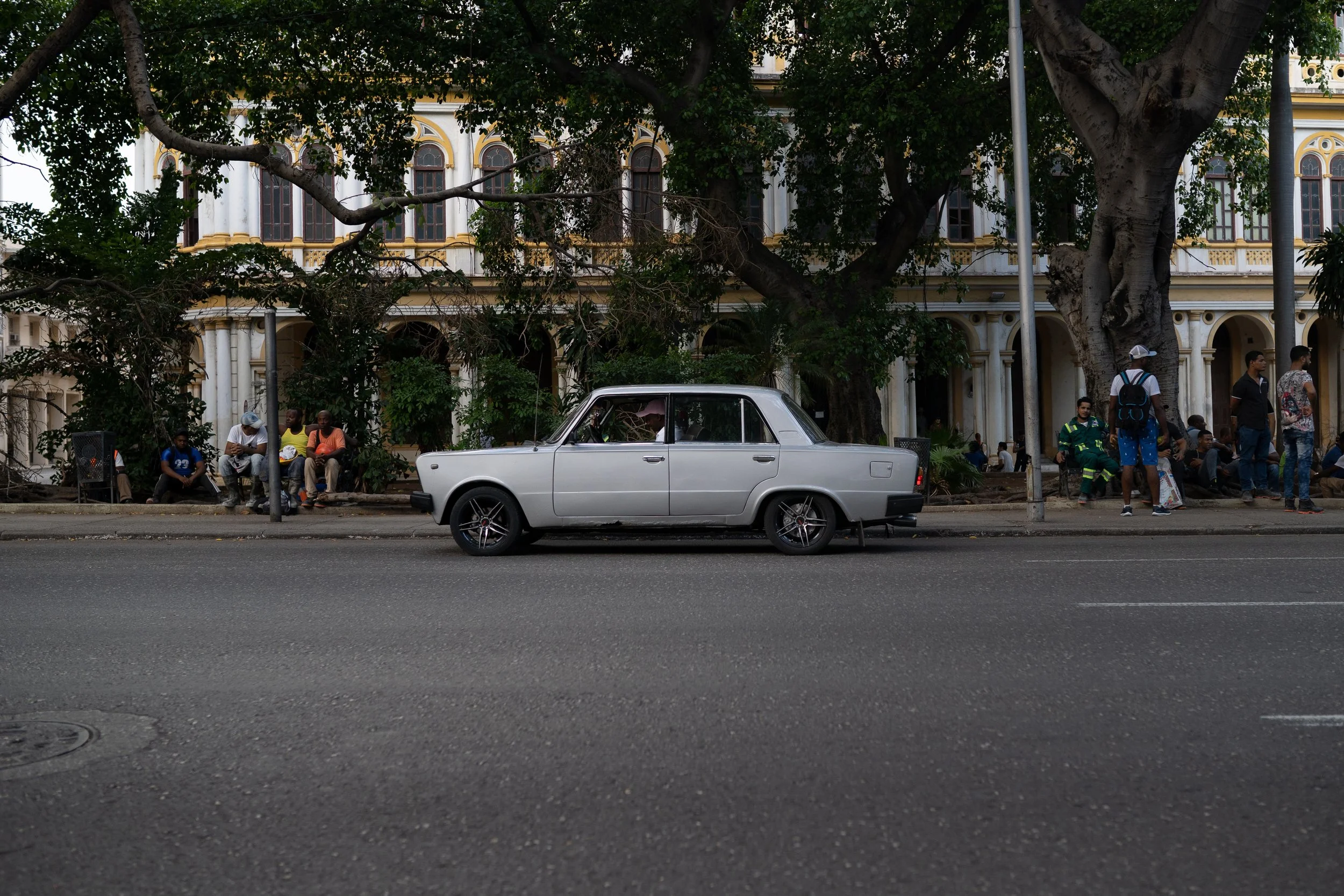 A gray four-door sedan parked on a city street with a large tree and colonial-style building in the background, several people sitting and walking nearby.