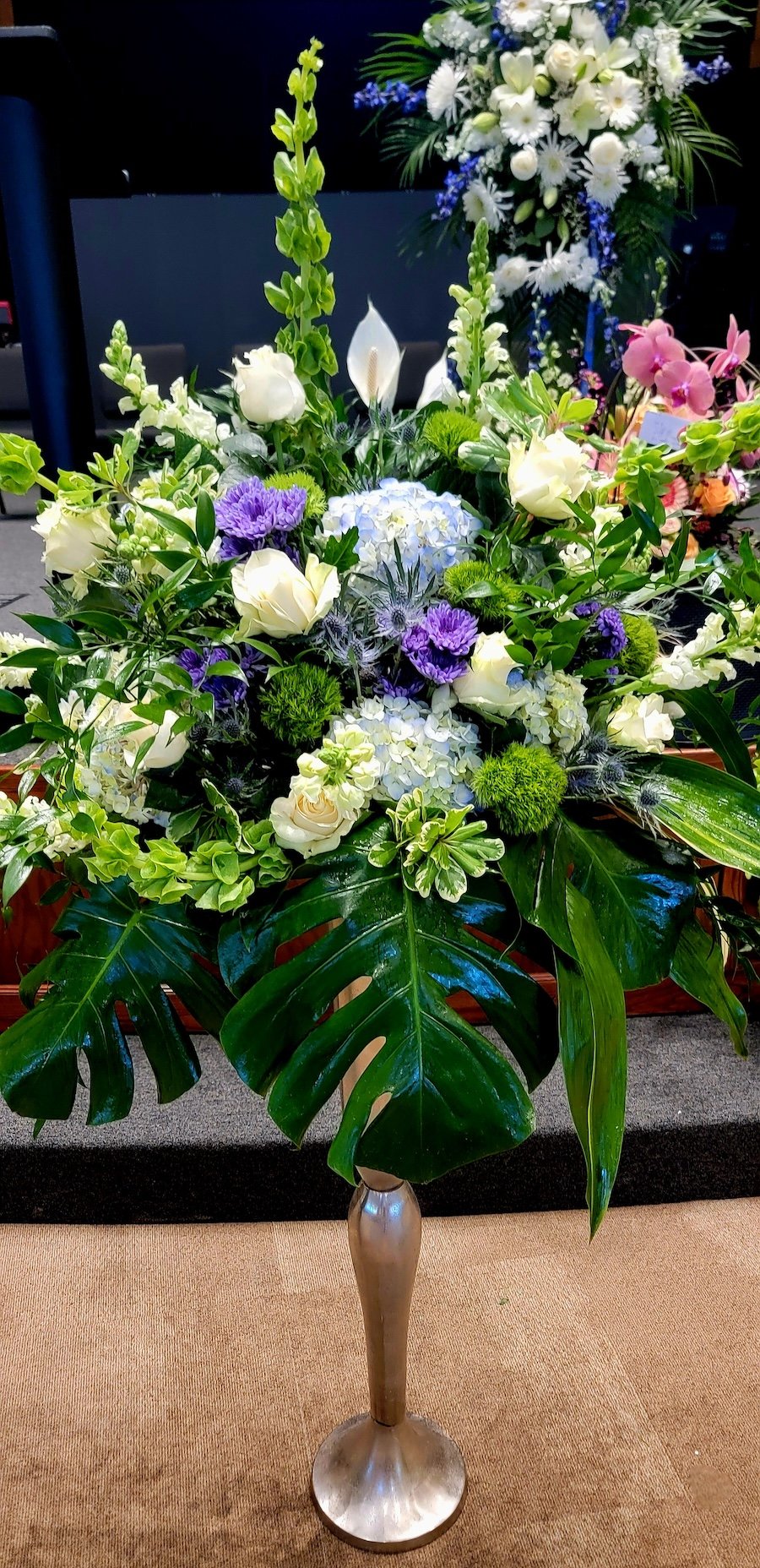 A tall floral arrangement with green leaves, white, purple, and pink flowers in a metallic gold vase.