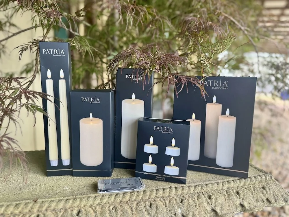 Various boxed led candles and light fixtures from Patria illumination arranged outdoors on a textured stone surface, some with artificial flames.
