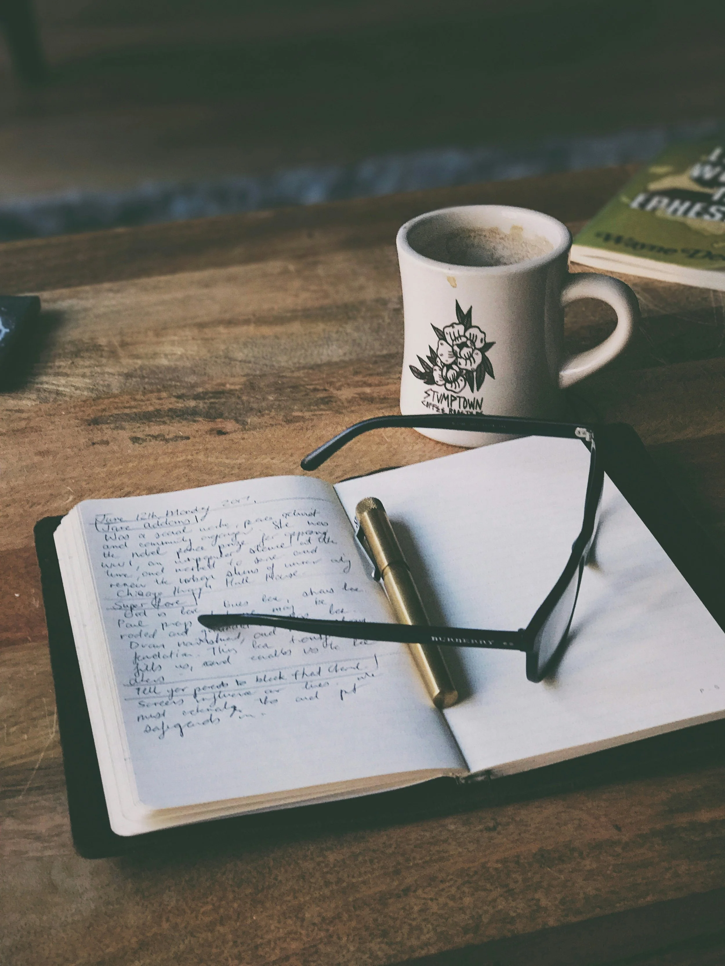 An image of items on a wood desk - a notebook with handwriting in pen on one page, a pen and glasses sitting on the notebook, with an empty stained coffee cup behind it.