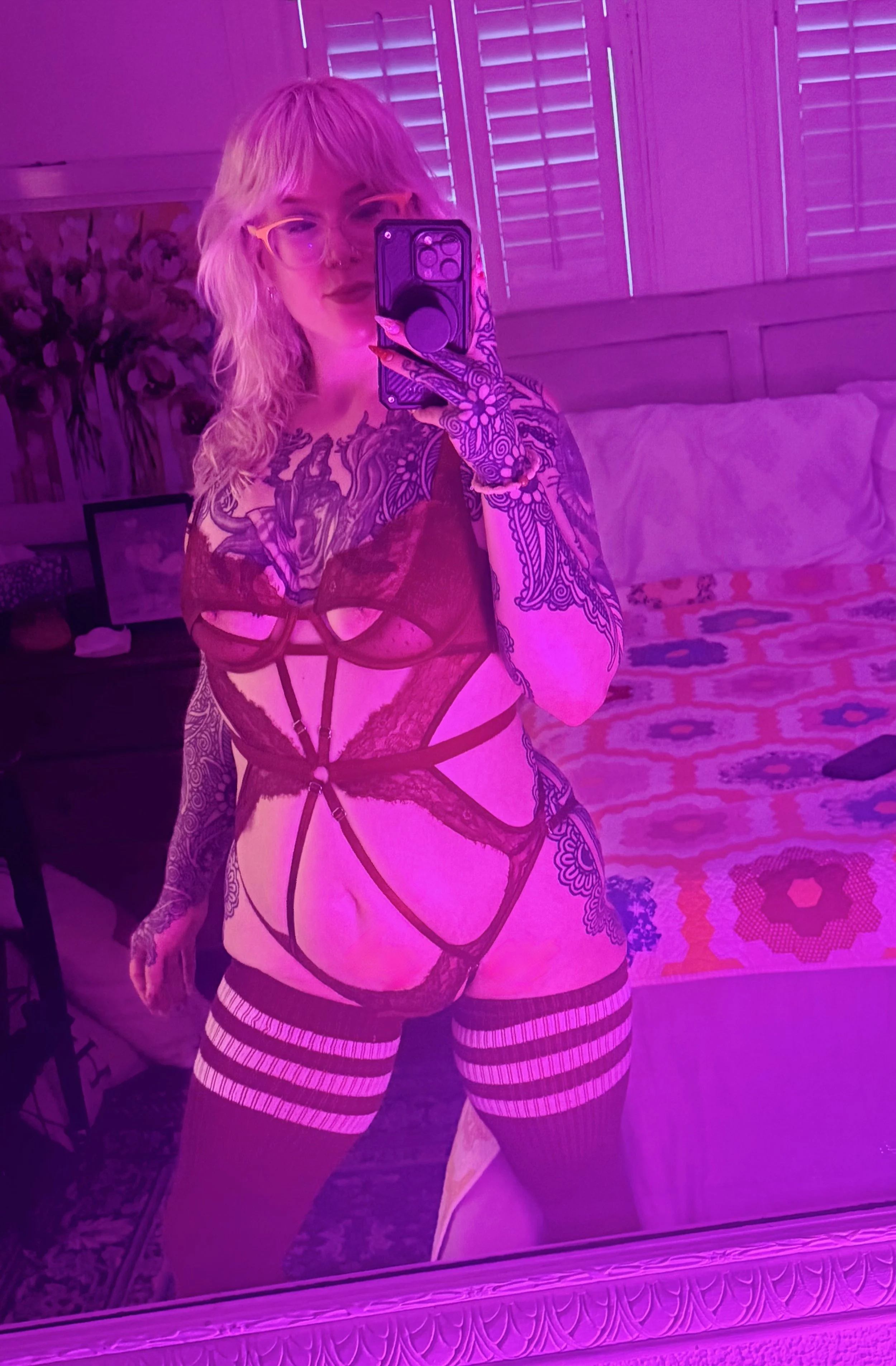 A woman taking a selfie in a mirror with purple lighting, wearing lingerie, thigh-high striped stockings, and showing tattoos on her arms and torso.