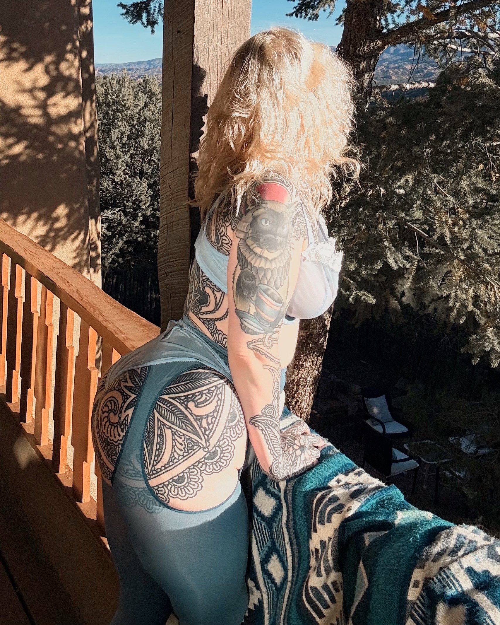 A woman with blonde curly hair and extensive tattoos is leaning on a wooden railing outdoors, looking away from the camera. She is wearing teal leggings and a white top, with a patterned blanket over the railing. The background features trees and a clear sky.