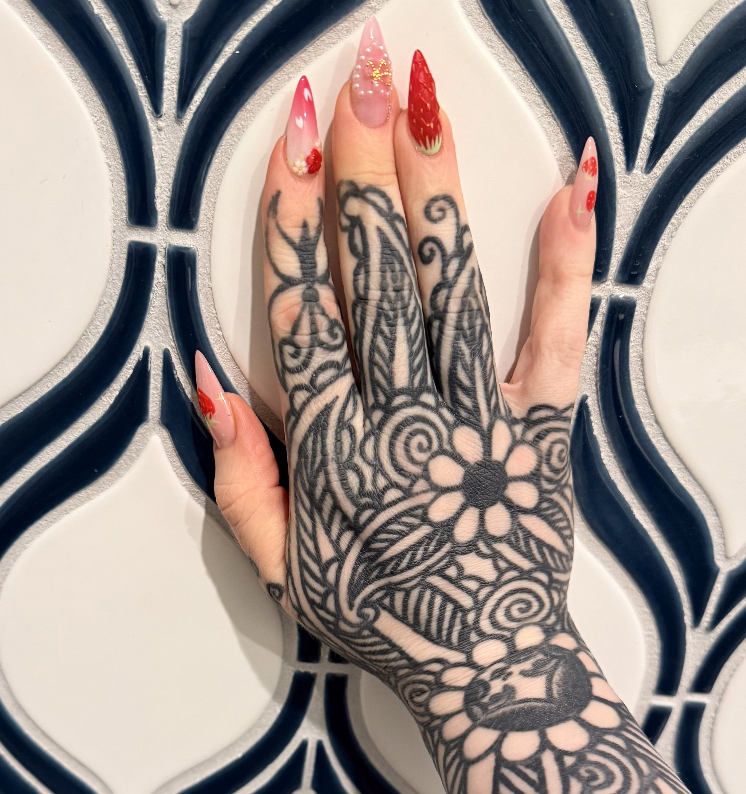 A tattooed hand with colorful nail art resting on a black and white decorative surface.