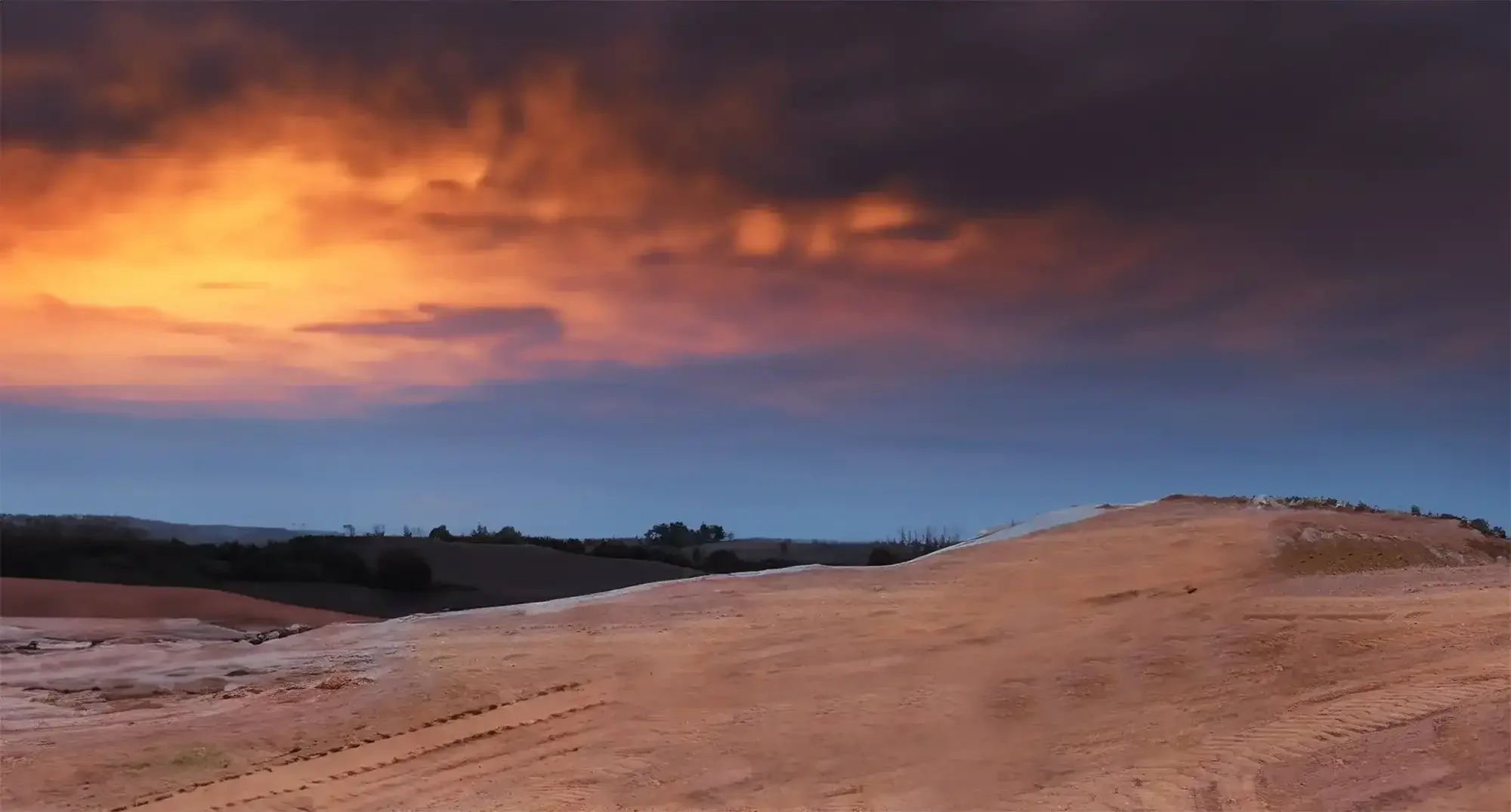 A landscape with rolling hills and a dramatic sky at sunset, featuring orange and dark clouds.