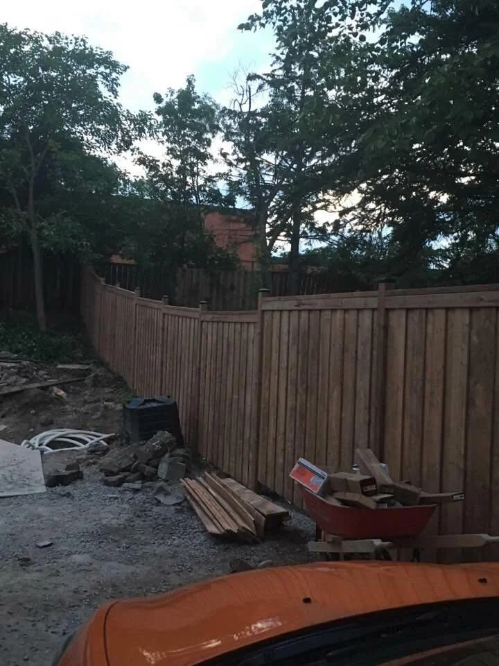 A wooden fence in a backyard with scattered construction materials, piles of lumber, and a red wheelbarrow filled with wood. The area is surrounded by trees.