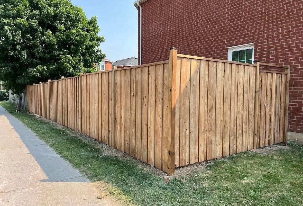 Wooden fence along a sidewalk next to a brick building and a tree.