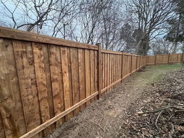 Long wooden fence with vertical planks, surrounded by trees and bare branches, on grassy and leaf-covered ground.