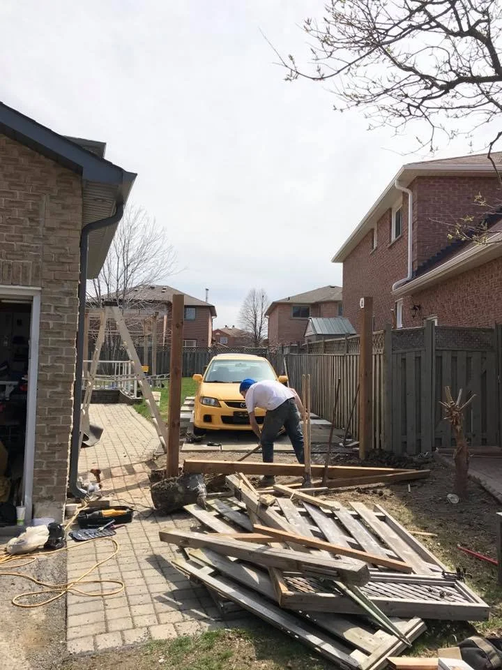 Man working on a backyard fence with a pile of wood nearby, a yellow car parked behind him.