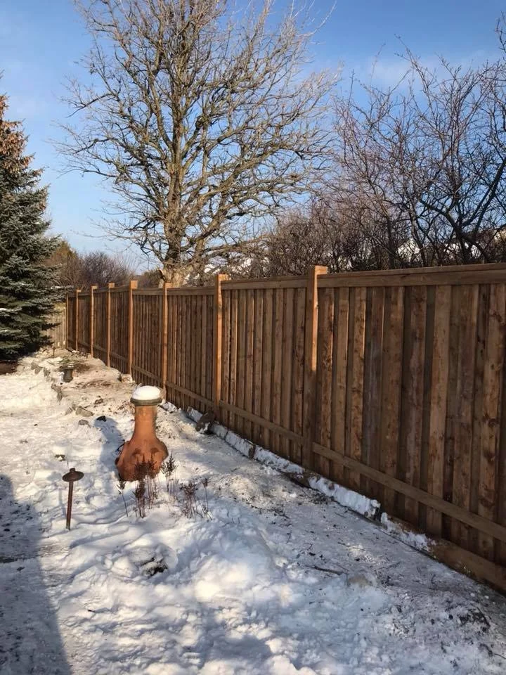 Snow-covered yard with wooden fence, leafless trees, and a clay chiminea.