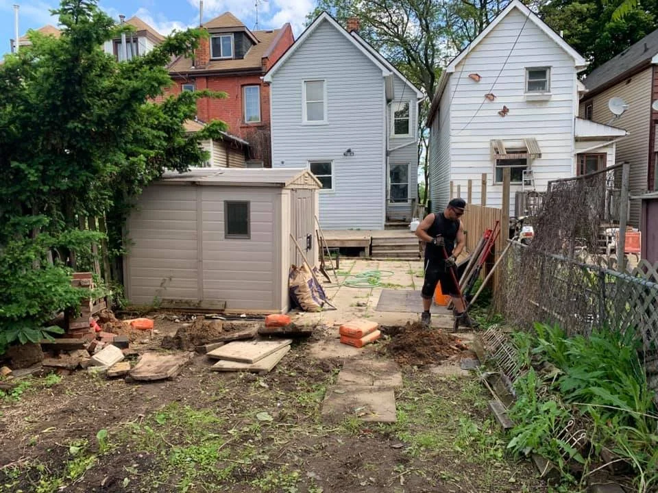 A backyard with a person working on the garden. There's a small shed, various tools, and building materials. Two houses are visible in the background. The ground appears disturbed, possibly indicating gardening or construction work.