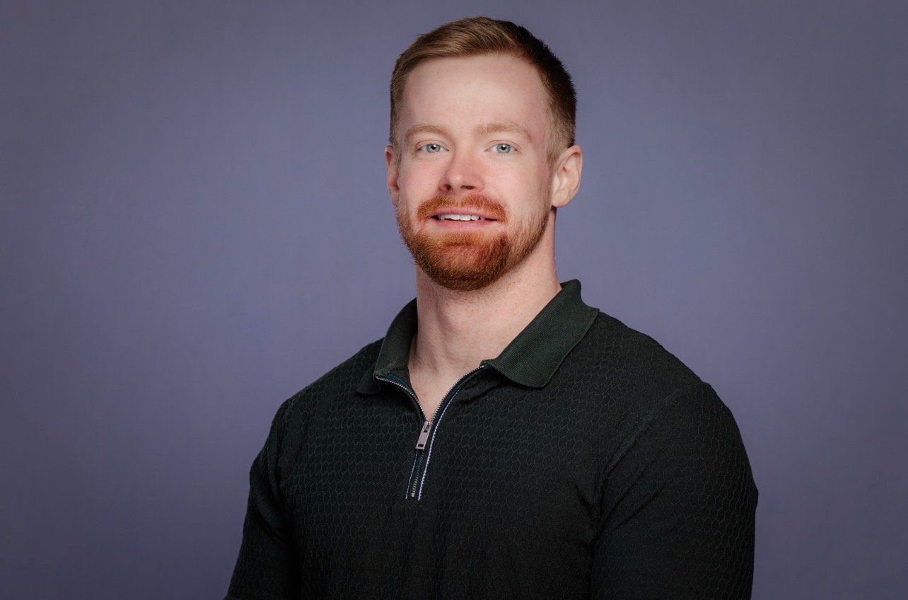 Portrait of a man with red hair and beard, wearing a black collared shirt, against a gray background.