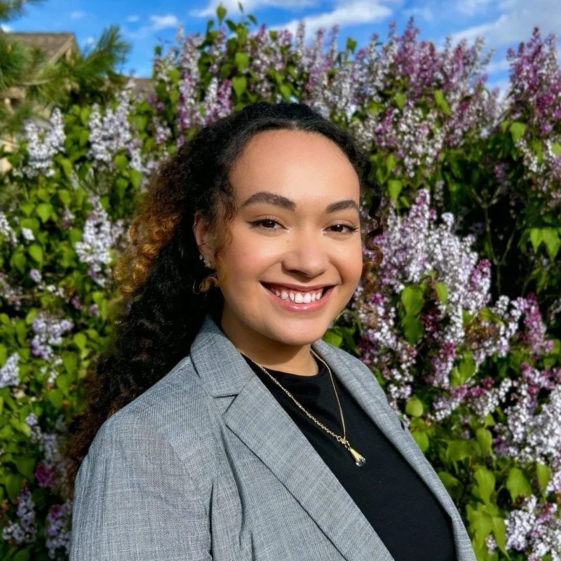 A young woman with curly hair and a blazer, smiling outdoors in front of a flowering bush with pink and white blossoms.