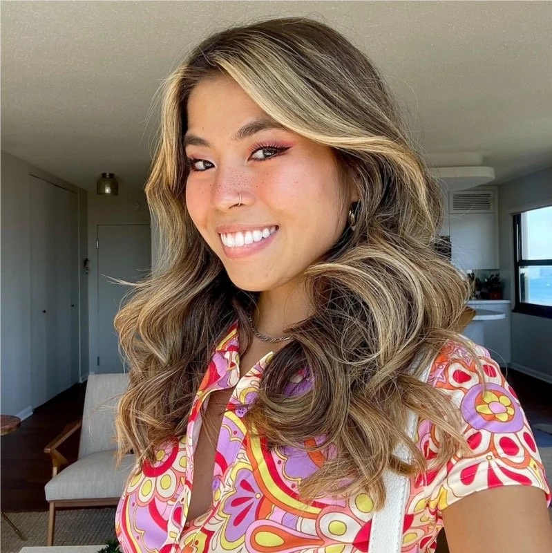 A young woman with long, wavy, light brown and blonde hair, smiling and winking. She is wearing a colorful patterned shirt with pink, purple, yellow, and red designs, and a silver chain necklace. The background shows a modern apartment with a chair, a door, and windows.