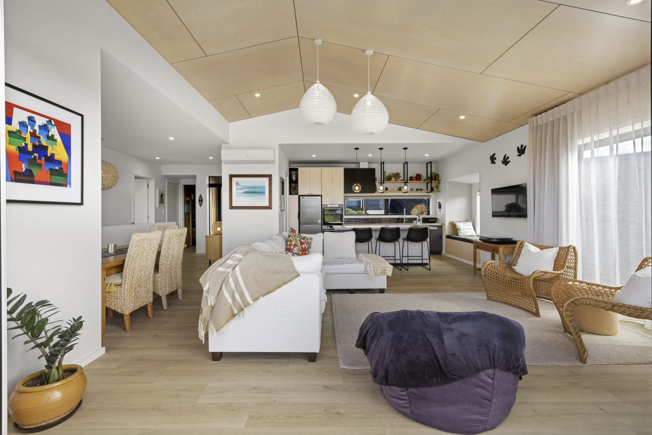 A spacious, modern living room with white walls, wooden floors, and a high, wooden ceiling. It features a white sofa with colorful pillows, a black bean bag chair, wicker chairs, and wall art. The open kitchen area has a breakfast bar with black chai