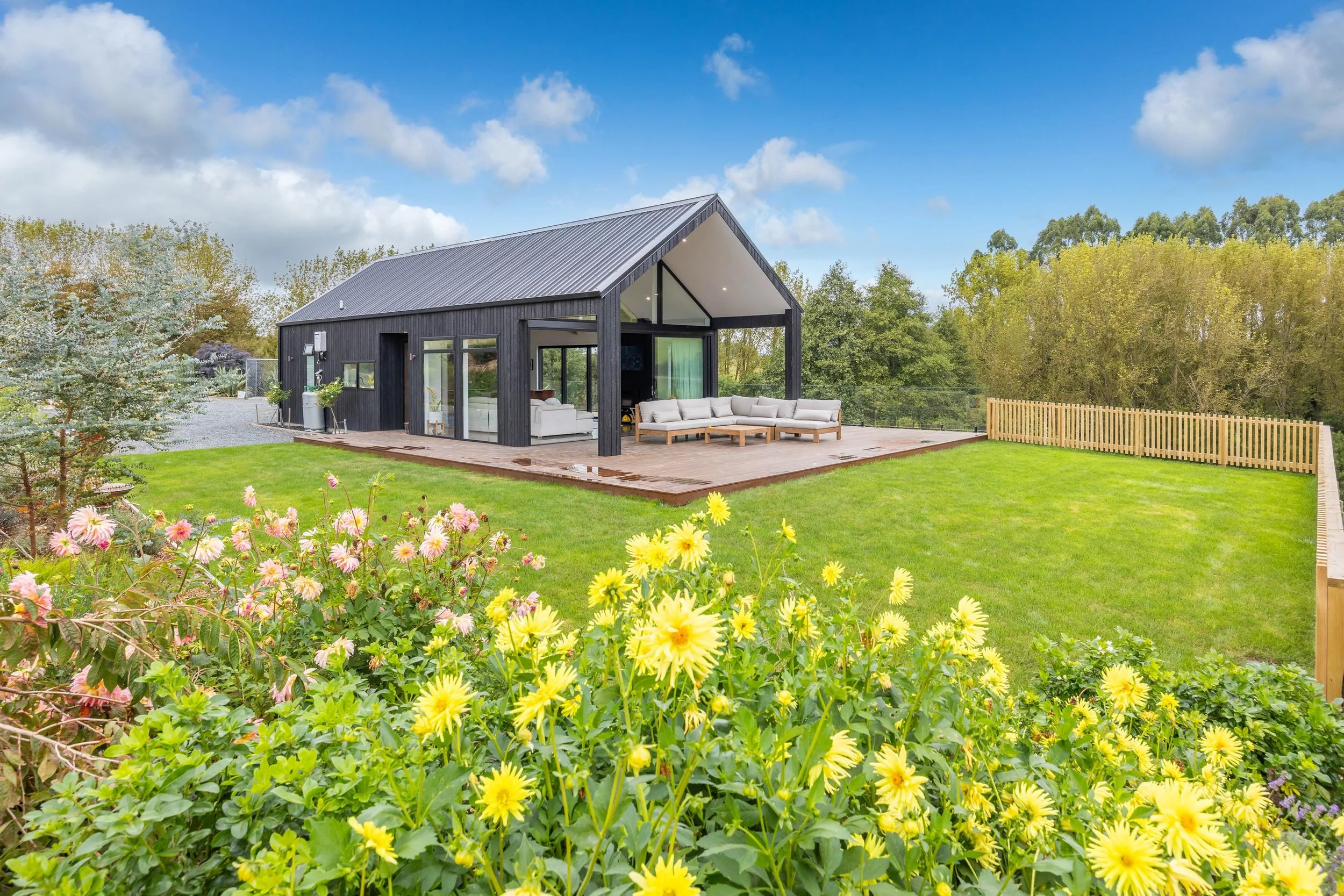 A modern black house with a sloped roof sits on a lush green lawn with yellow flowers in the foreground. The house has large glass windows and an outdoor seating area on a wooden deck, surrounded by trees and a blue sky with white clouds.