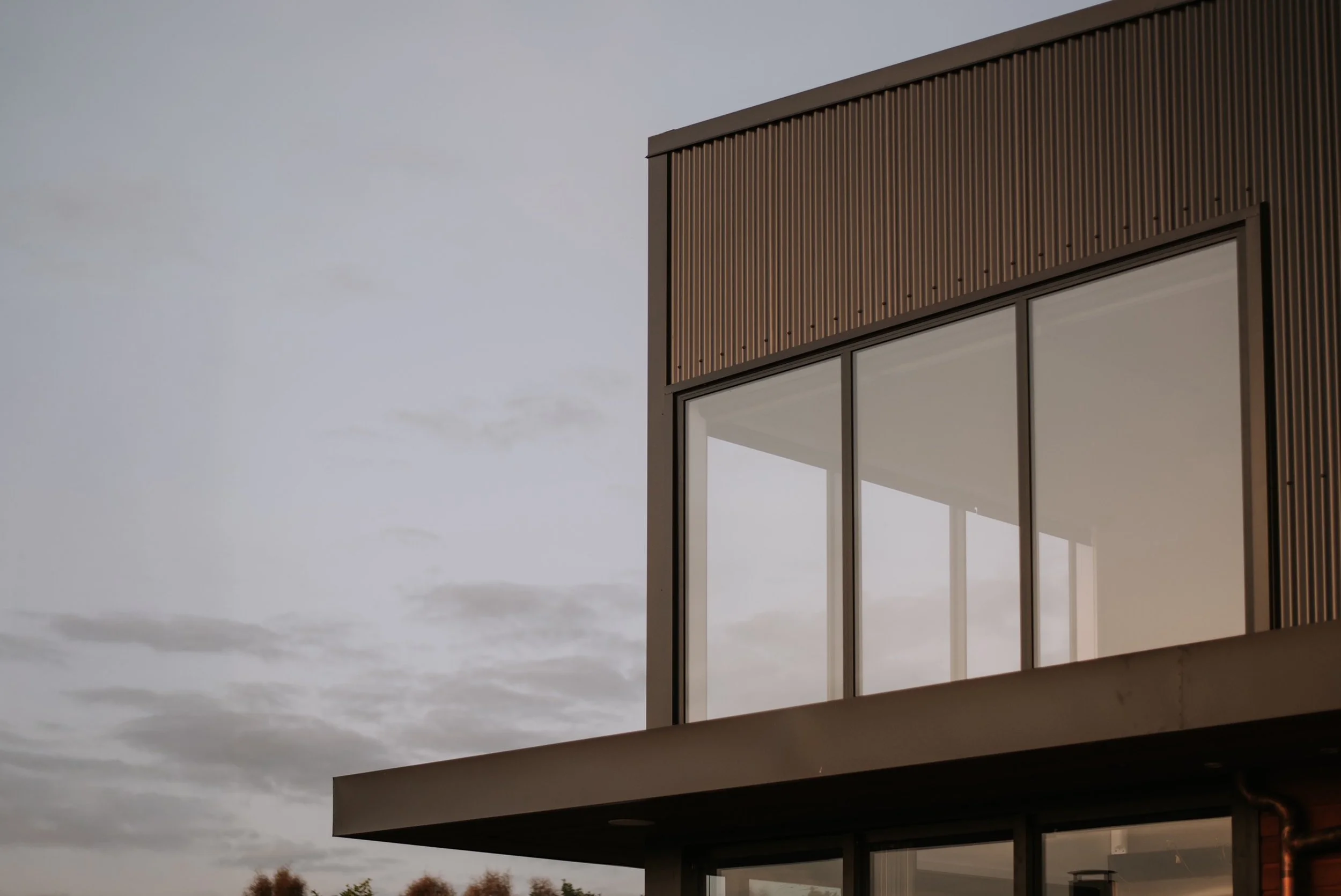 Part of a modern building with a flat roof, large glass windows, and brown siding, against a cloudy sky.
