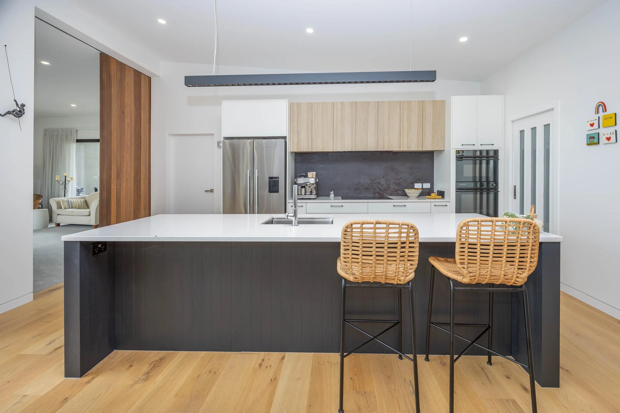 Modern kitchen with white countertops, wooden cabinets, stainless steel refrigerator, black backsplash, and two woven rattan bar stools at the counter, with a living room area in the background.