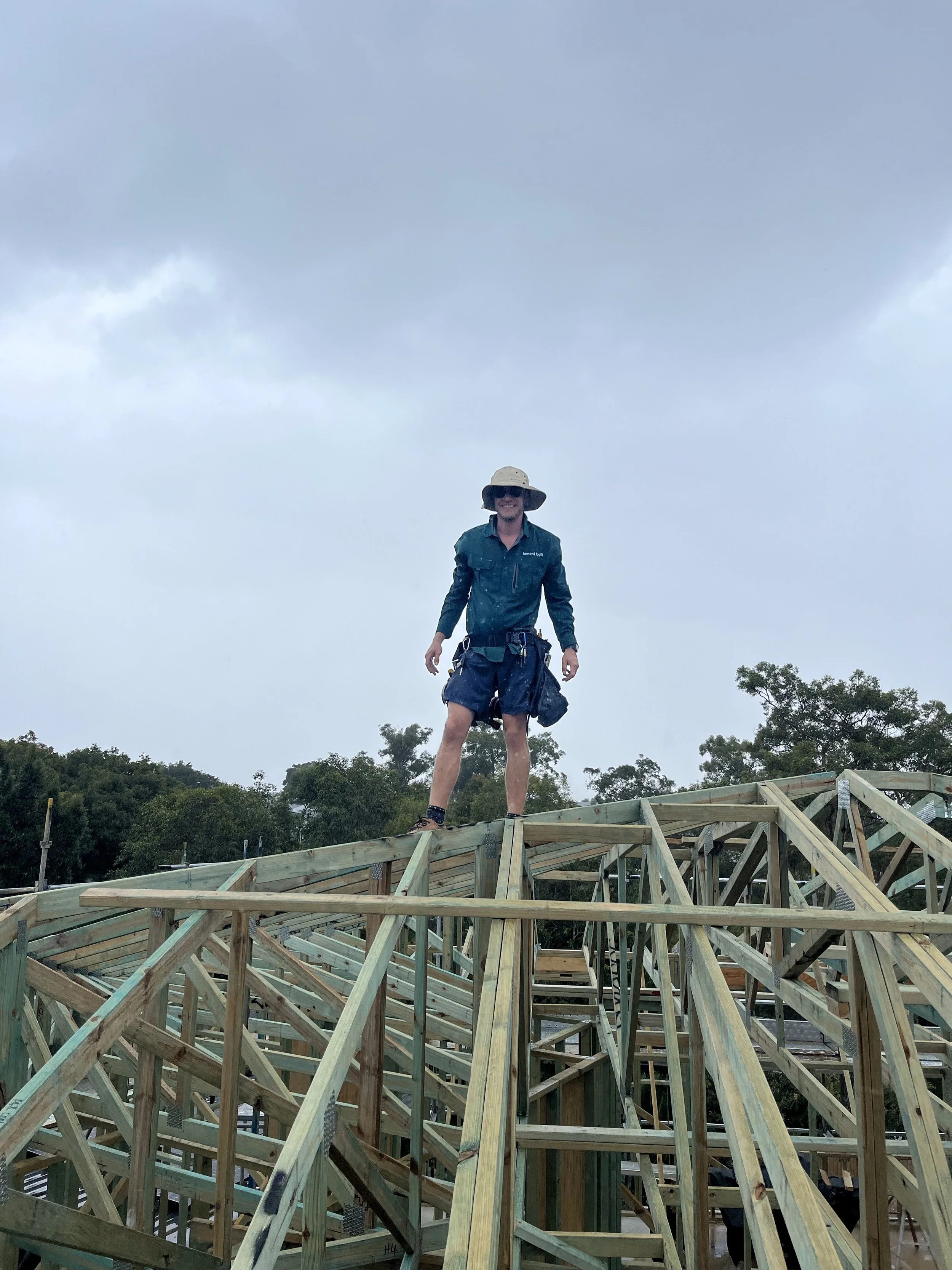 A construction worker in a hat stands on the wooden frame of a building under a cloudy sky.