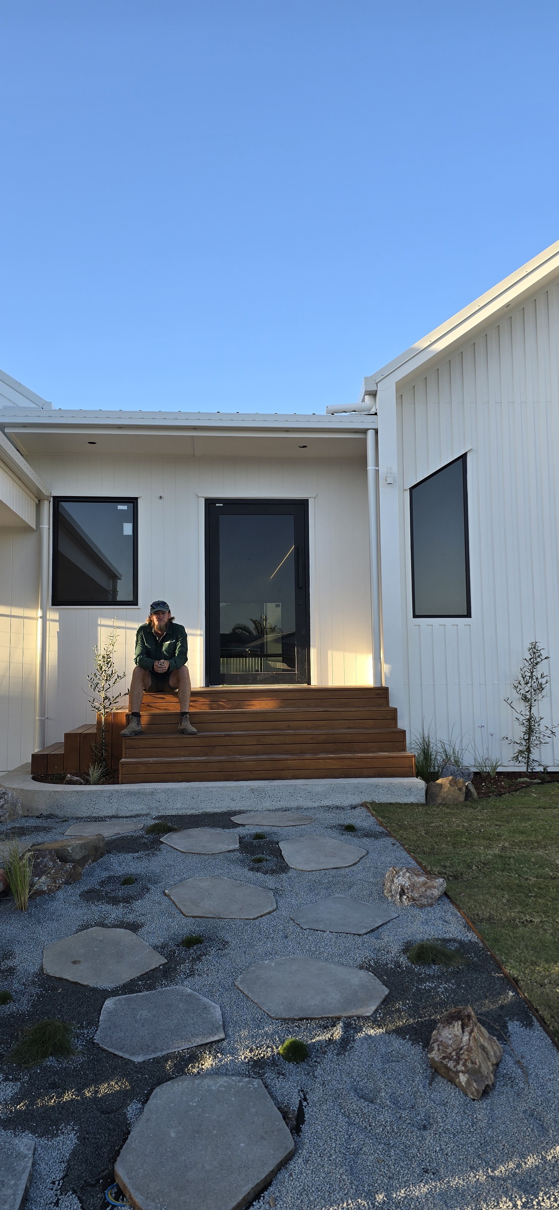 A person sitting on wooden stairs outside a modern white house with large windows and a glass door, under a clear blue sky.