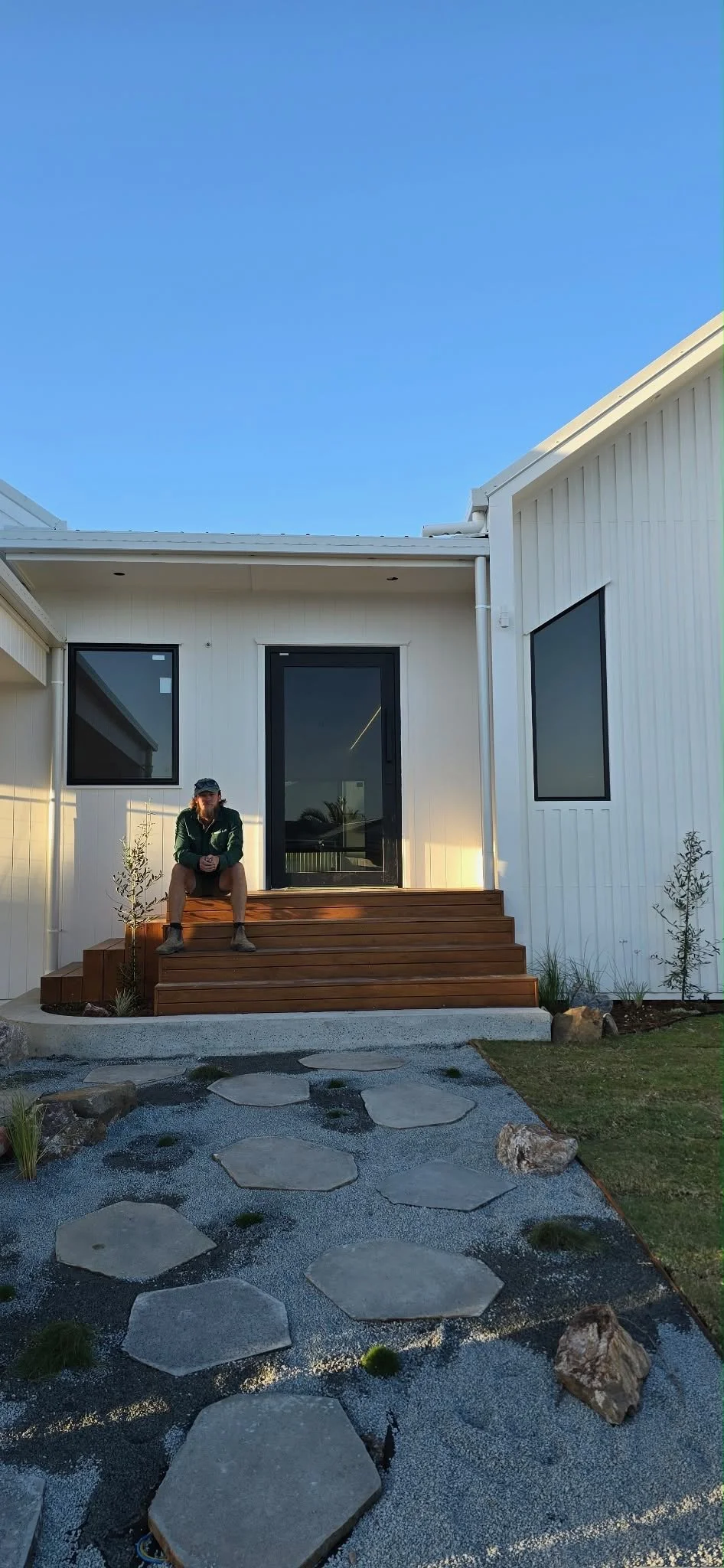 Person sitting on wooden steps outside a modern white house with black-framed windows and door, small trees, rocks, and a pathway with stepping stones under a clear blue sky.