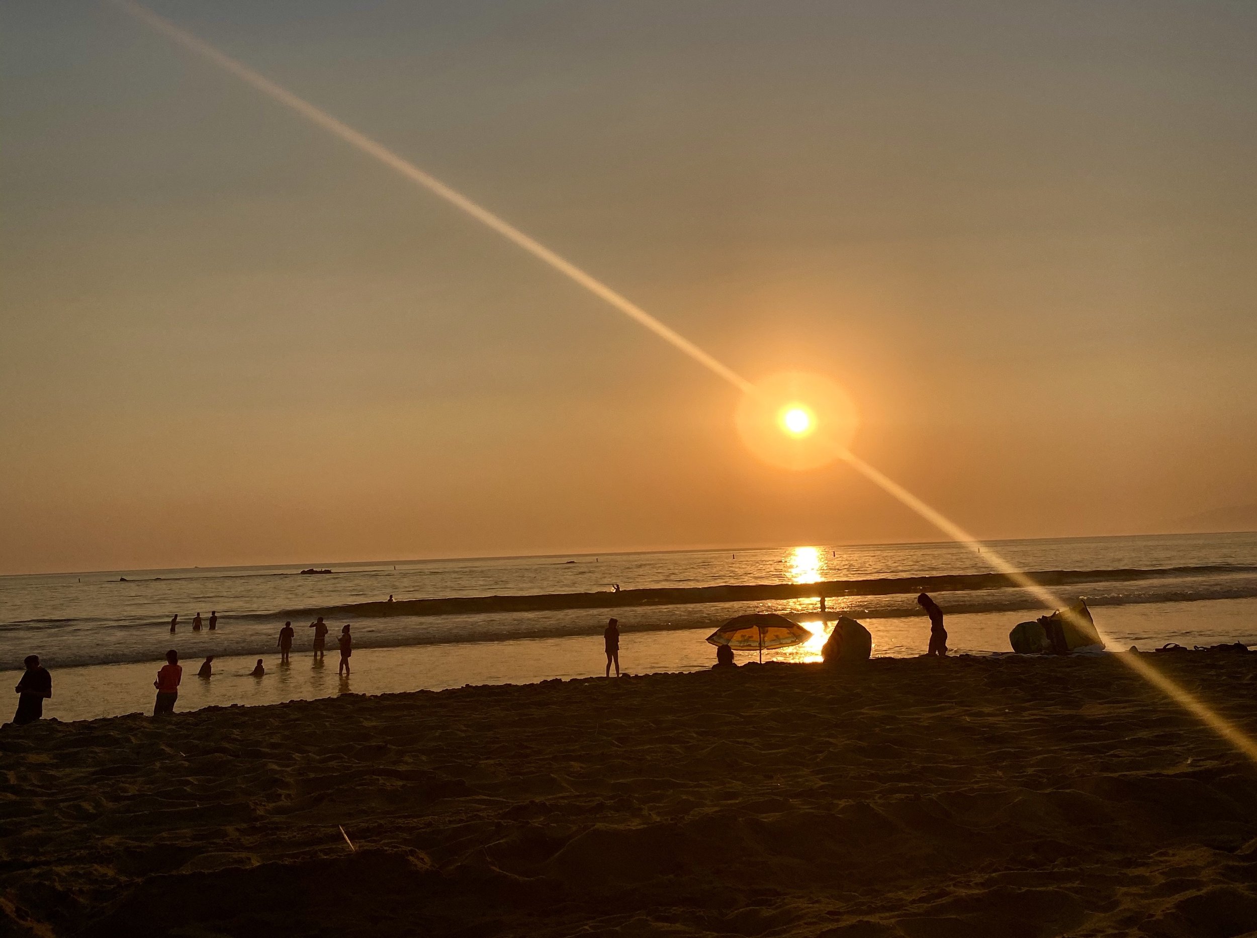 Beach at sunset with people silhouetted against the ocean and a visible sunbeam across the sky.