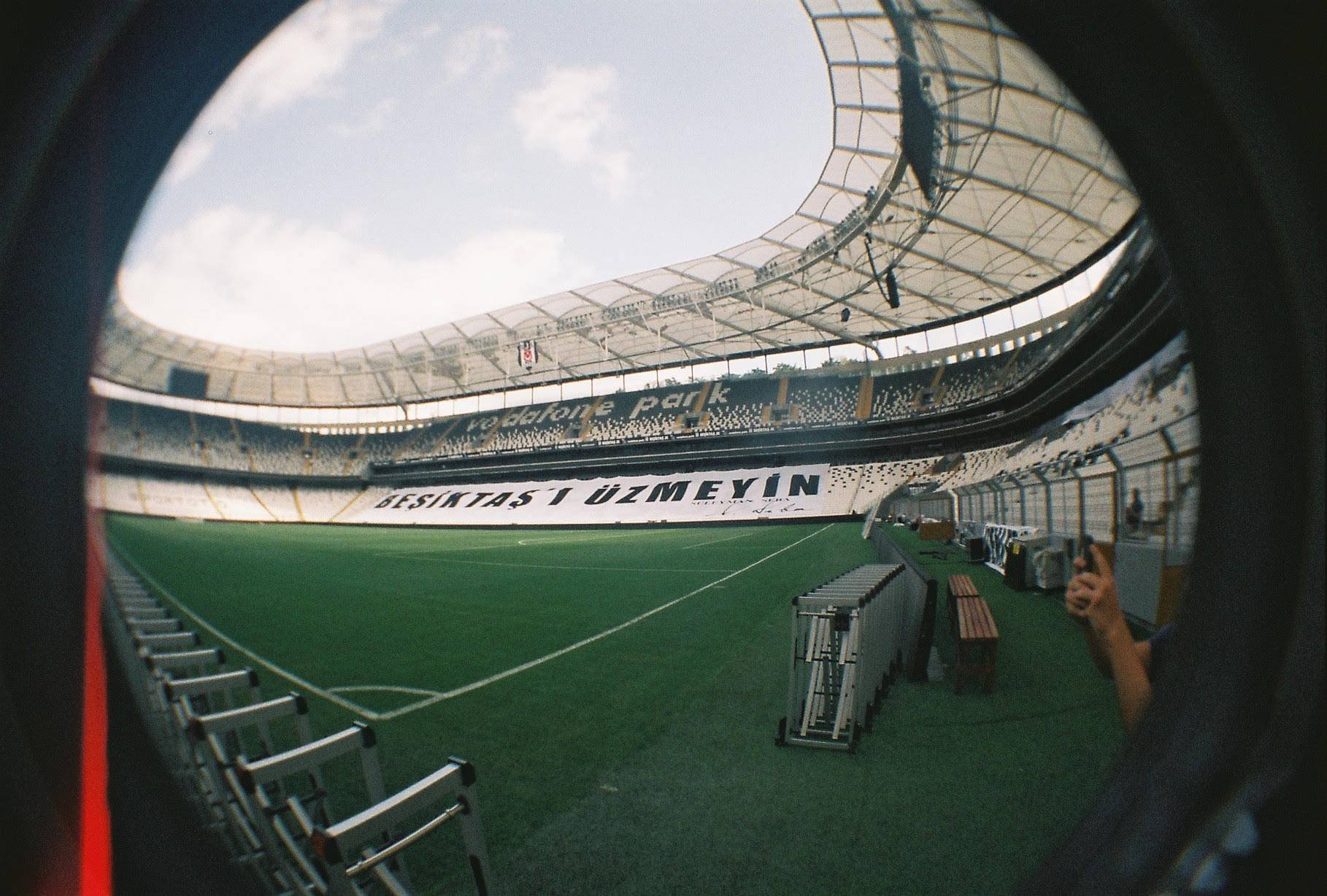 Wide-angle view of a football stadium with green pitch, empty stands, and a large banner displaying text. The stadium is covered with a partial roof.