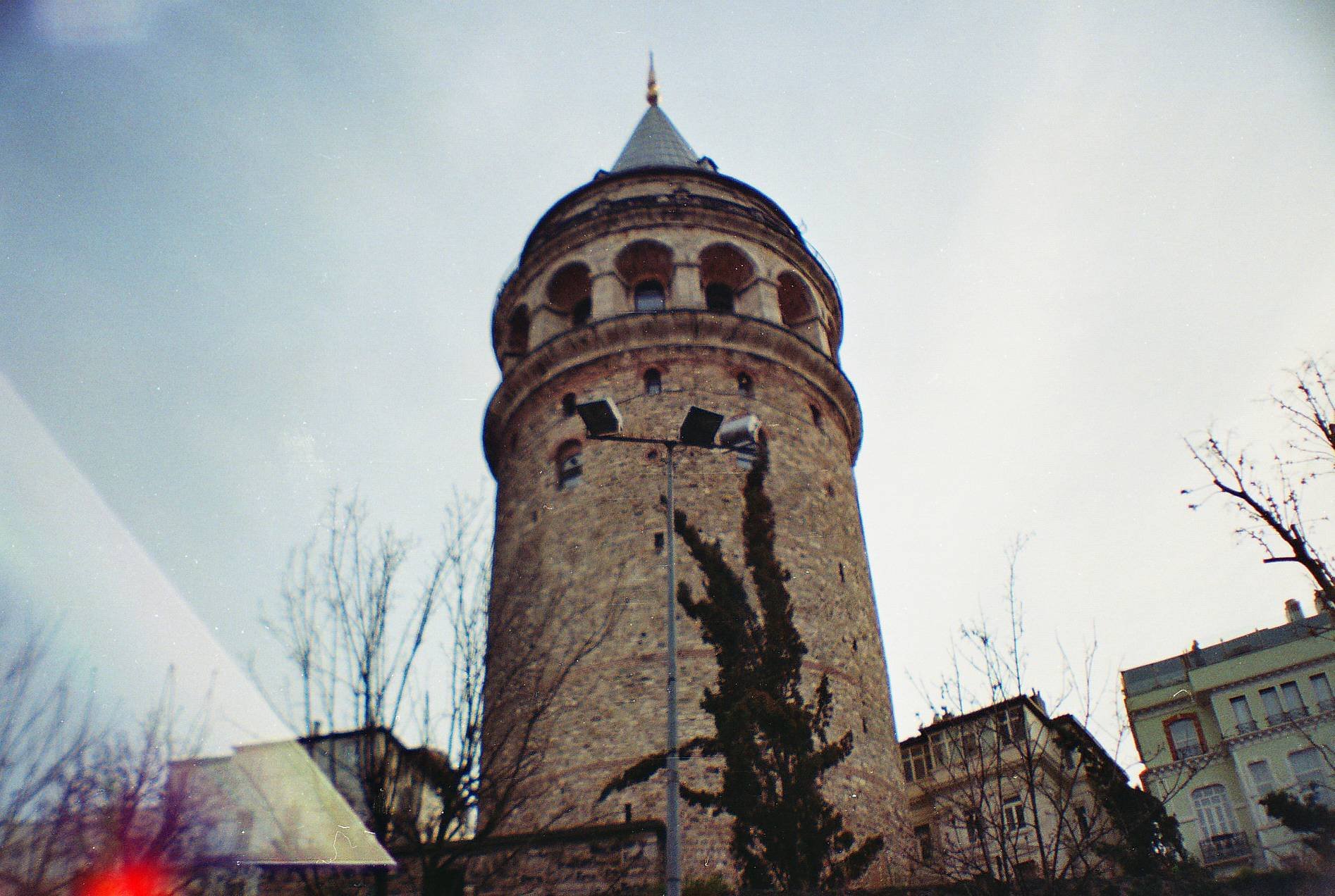 Historical tower with conical roof and round windows, surrounded by buildings and leafless trees, against a clear sky.