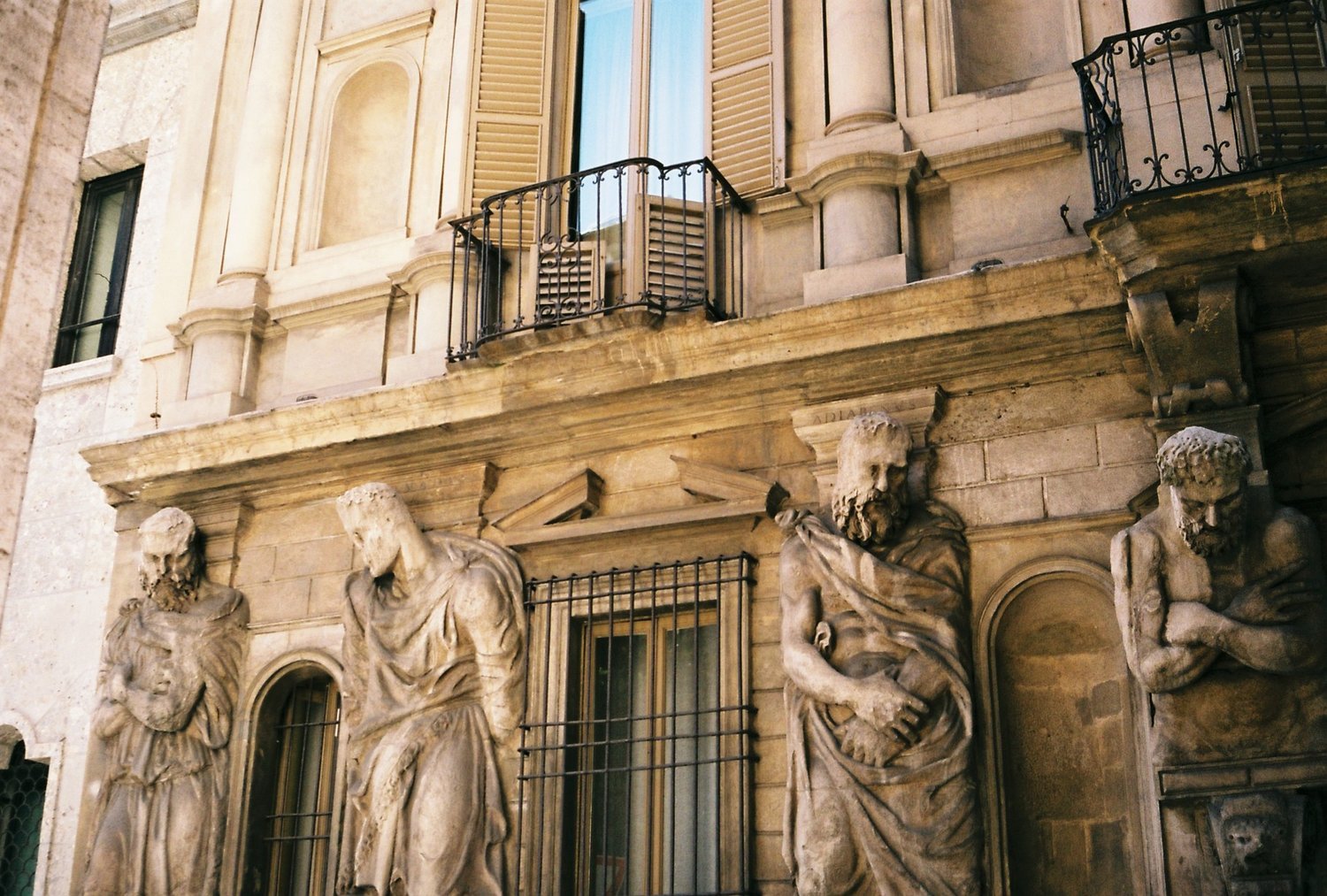 Historic building facade with four stone statues of robed figures, balcony above, and architectural details in warm tones.