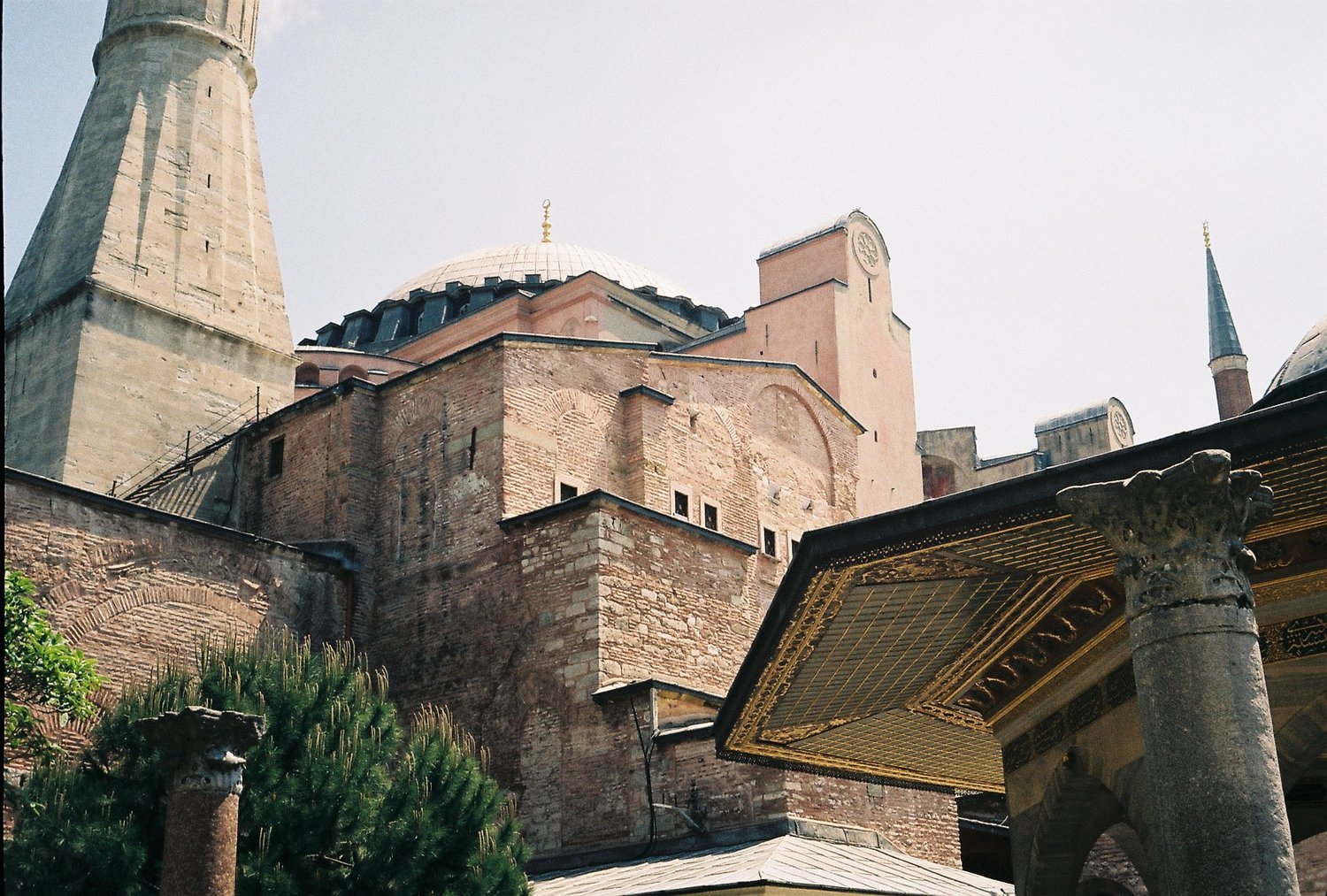 Exterior view of Hagia Sophia showcasing its domes and brick walls, with a minaret and an ornate columned structure in the foreground.