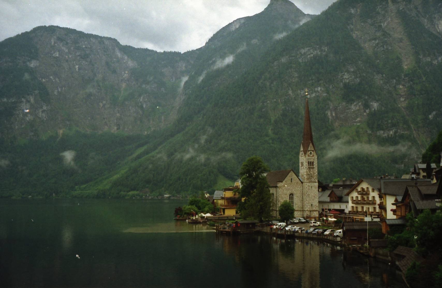 Scenic view of Hallstatt, Austria, featuring a church with a tall steeple by a lake, surrounded by mountains and greenery.