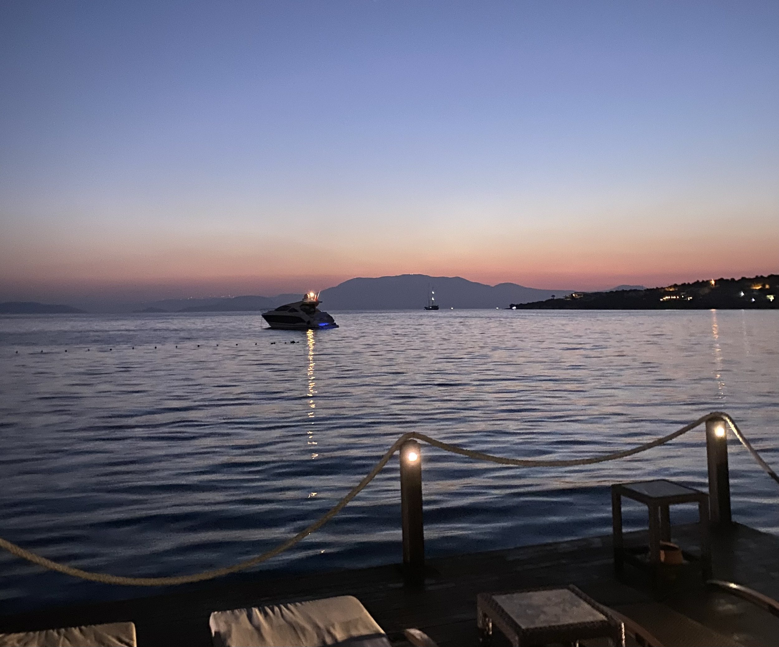 Evening view of a calm sea with a yacht and distant hills under a colorful sunset sky. Wooden deck with seating and rope railing in the foreground.