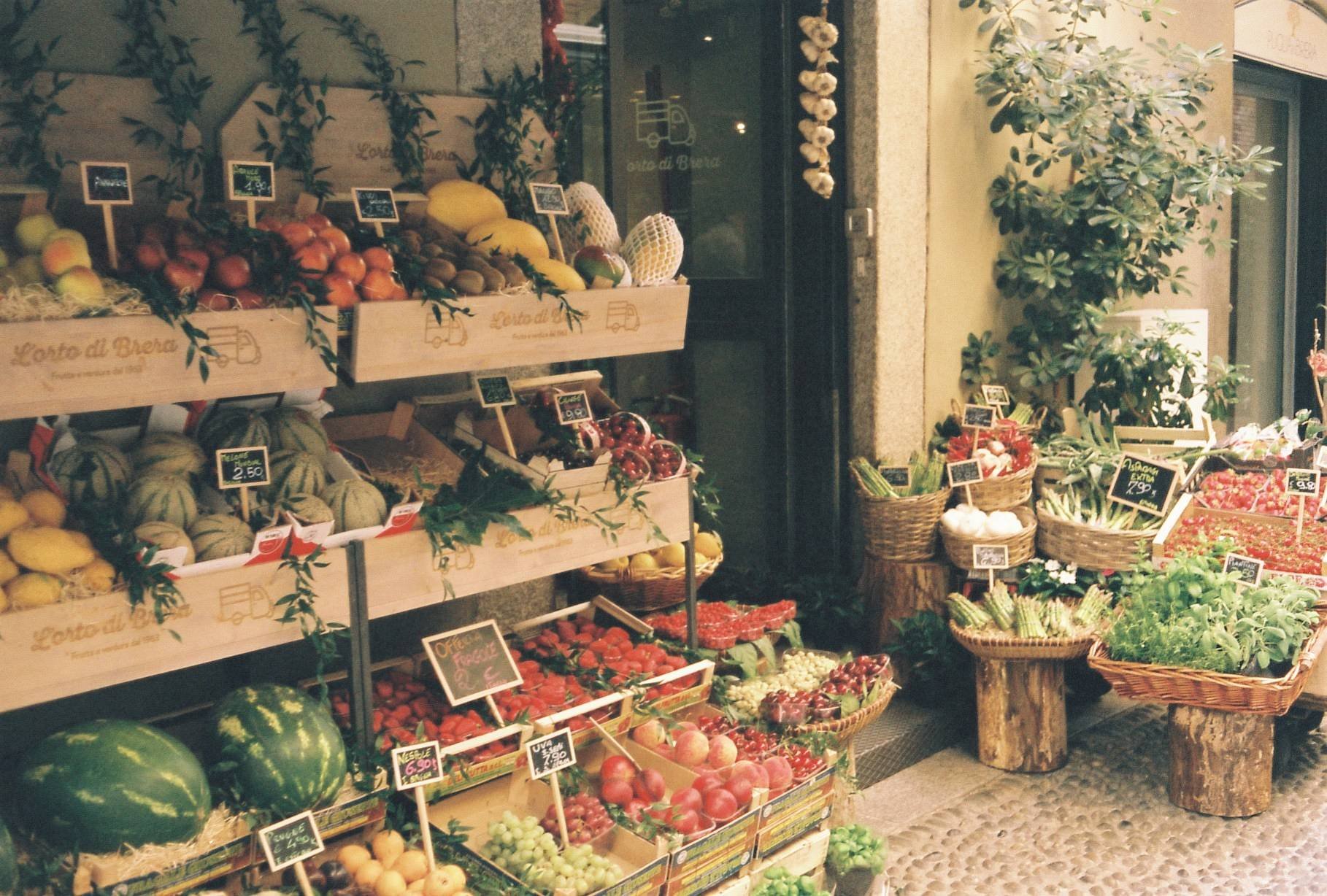 Outdoor market stall with various fruits and vegetables including watermelons, melons, tomatoes, grapes, and green vegetables, displayed in wooden crates and baskets.