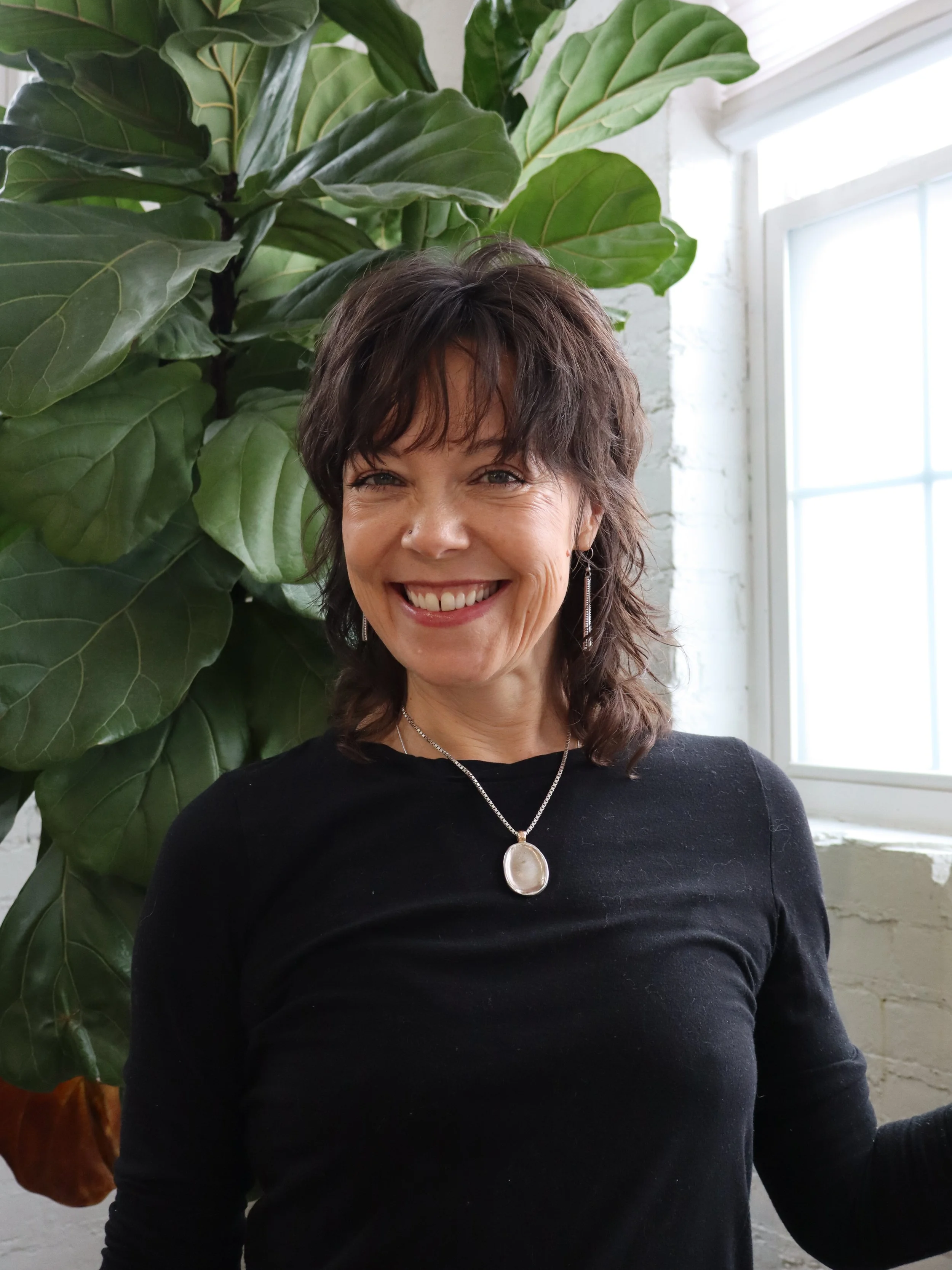 A woman with short, dark, wavy hair, smiling, wearing a black top and jewelry, standing near a large green plant indoors with a window in the background.