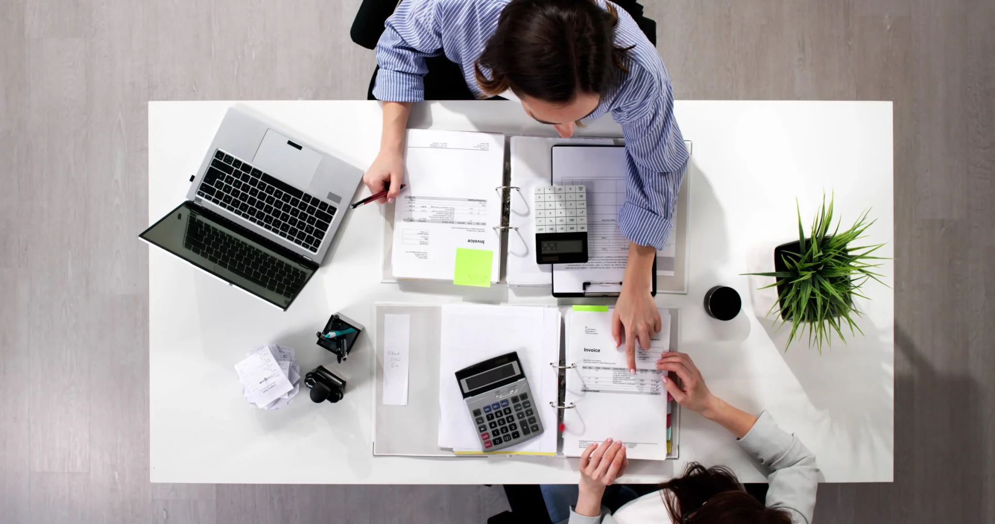 A top-down view of a workspace with two people working on financial documents. The workspace features a laptop, calculators, paperwork, a potted plant, and office supplies on a white desk.