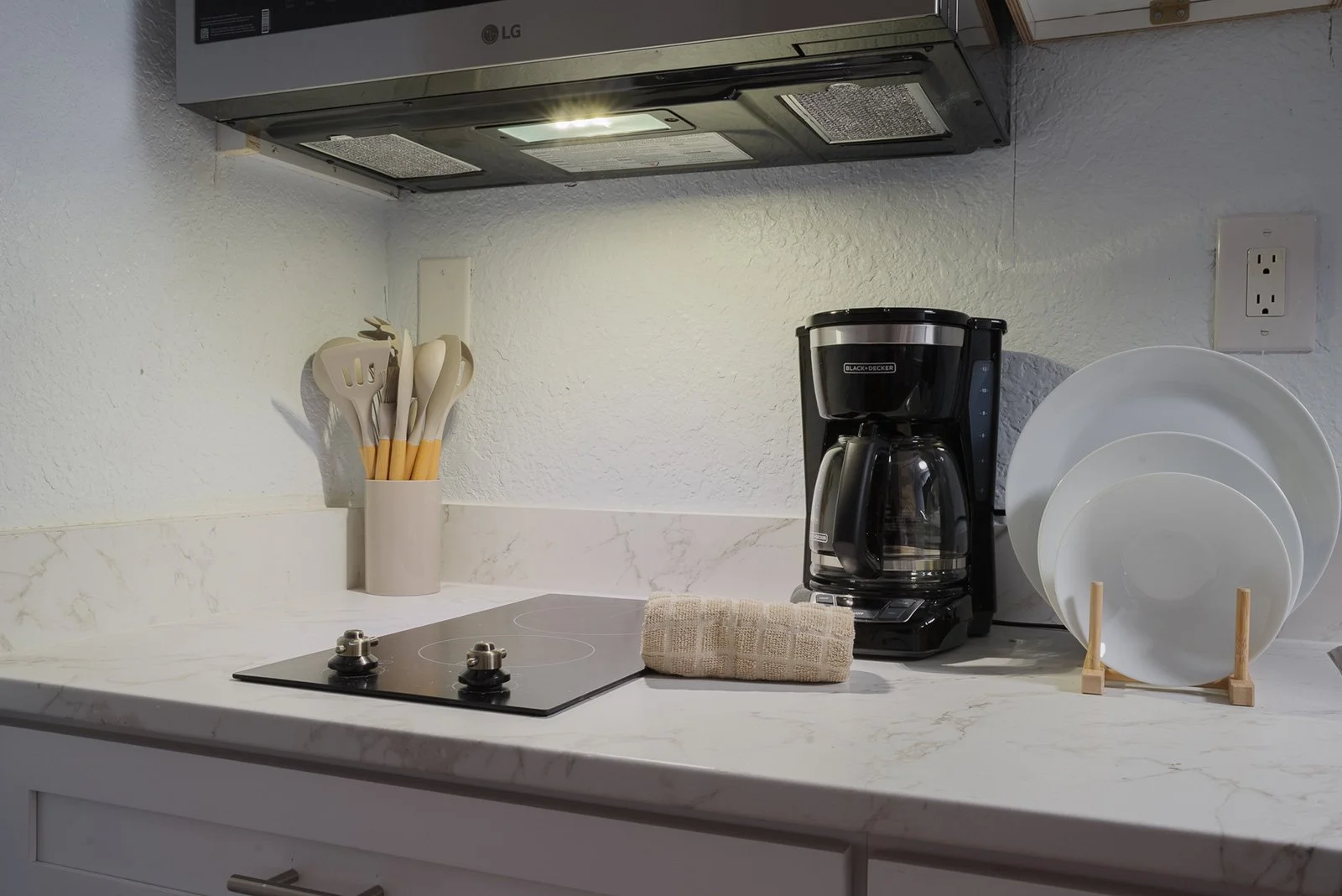 Kitchen countertop with a coffee maker, white plates on a wooden stand, a utensil holder with white and beige utensils, a folded towel, and an electric stovetop with two burners under a range hood.