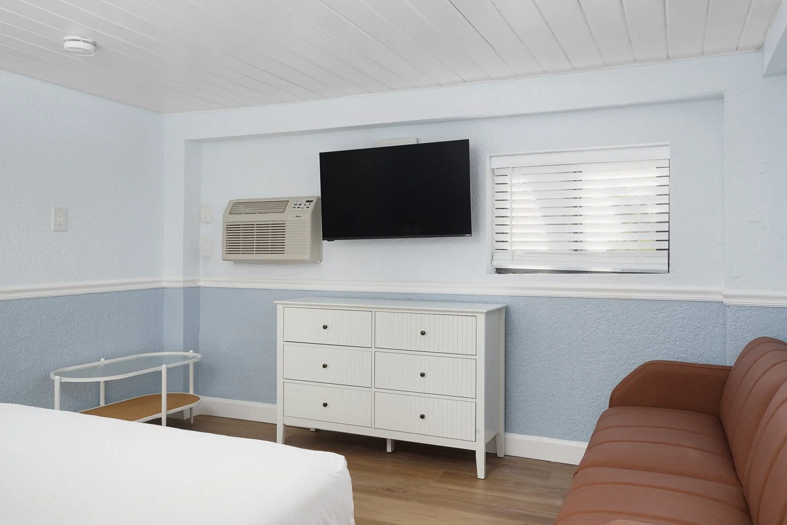 A room with light blue walls, a flat-screen TV on the wall, a window with blinds, an air conditioning unit, a white dresser, a brown sofa, and part of a white bed in the foreground.