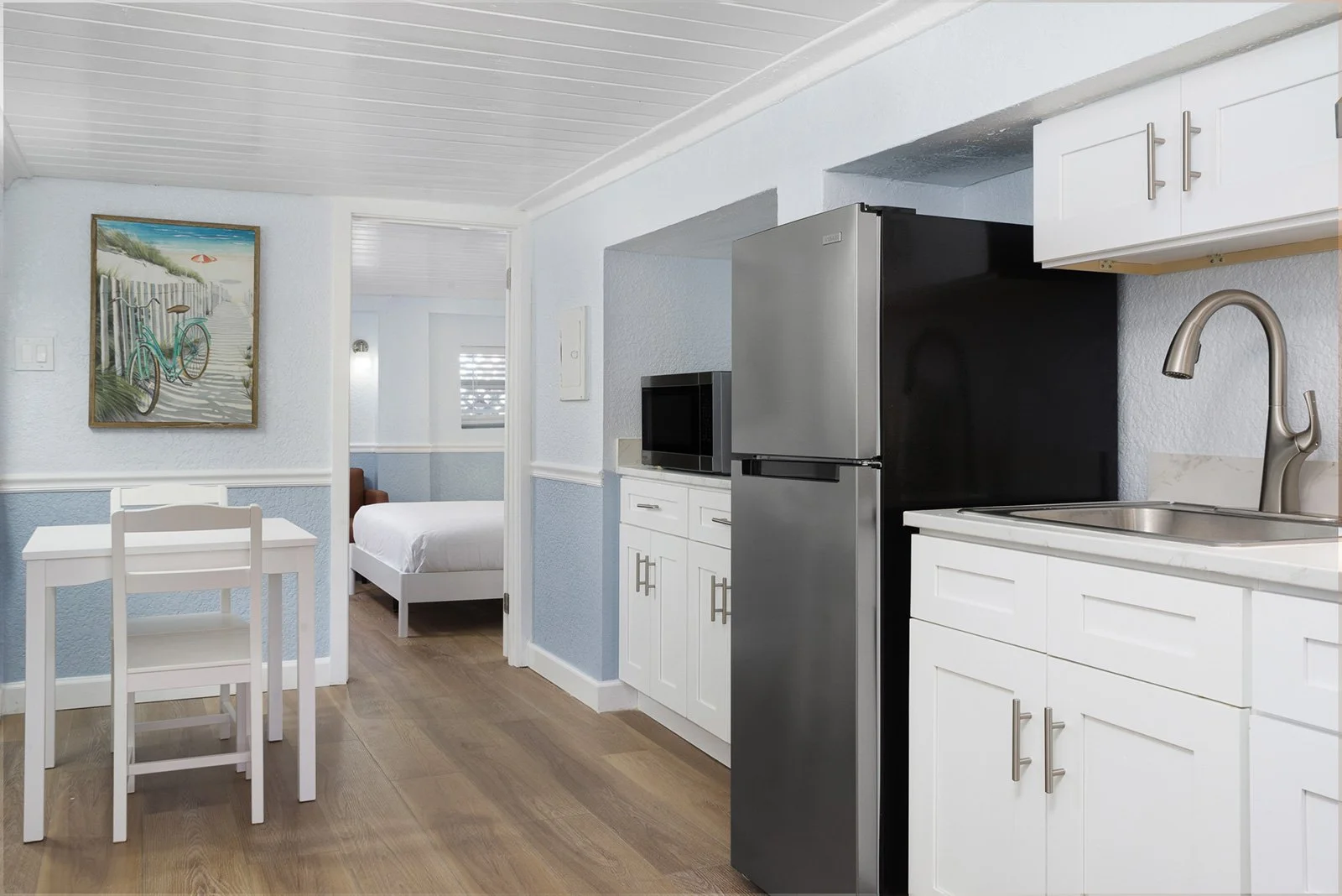 A beach boutique hotel room with a cozy kitchen with white cabinets, stainless steel refrigerator, microwave, sink, and a small dining table with two chairs. A door leads to a bedroom with a white bed and a painting of a bicycle and beach scene on th