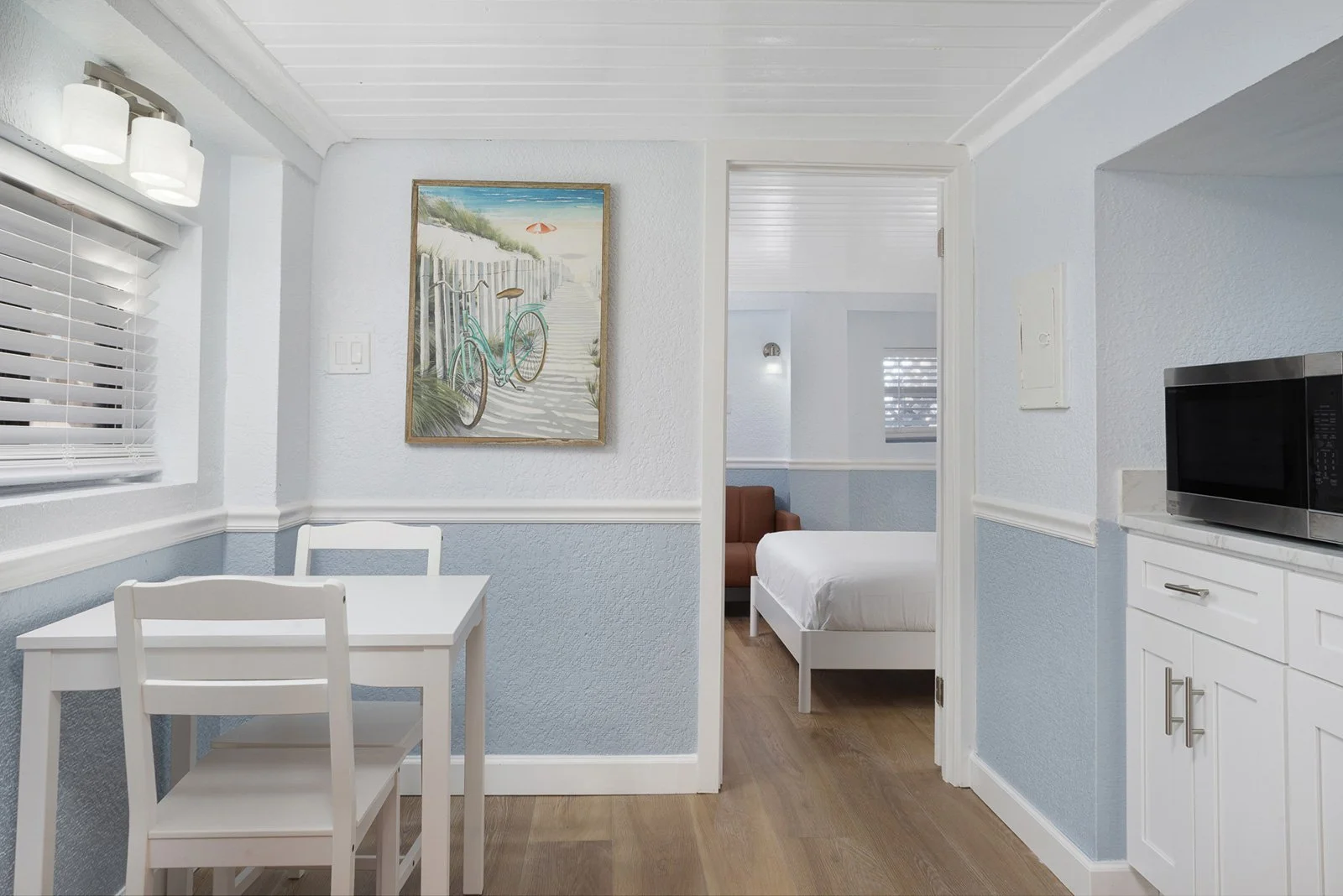 A cozy, brightly lit beach boutique hotel bedroom with light blue and white walls, wooden flooring, a white desk with matching chairs, a wall art of a bicycle and beach scene, a small kitchenette with a microwave, and a view into the adjacent bedroom