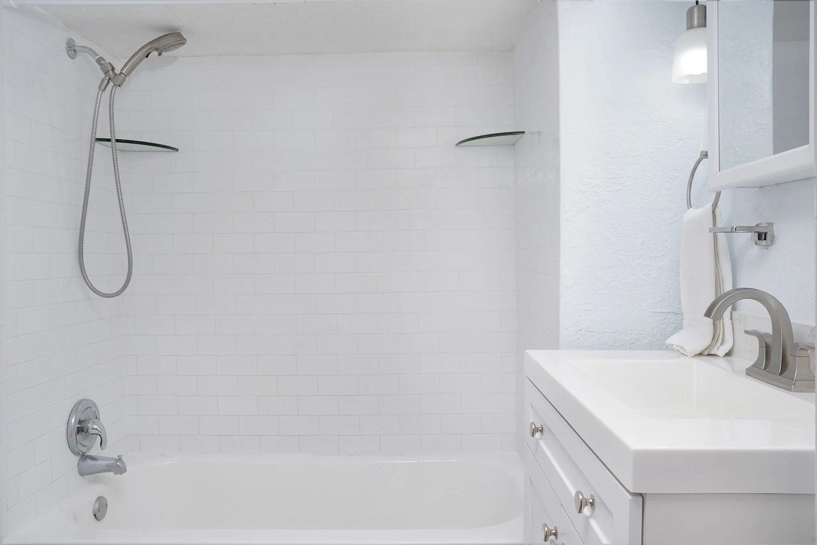 Clean white bathroom with bathtub and shower, small glass shelves, white vanity with silver knobs, silver faucet, towel rack with towel, mirror, wall-mounted light fixture.