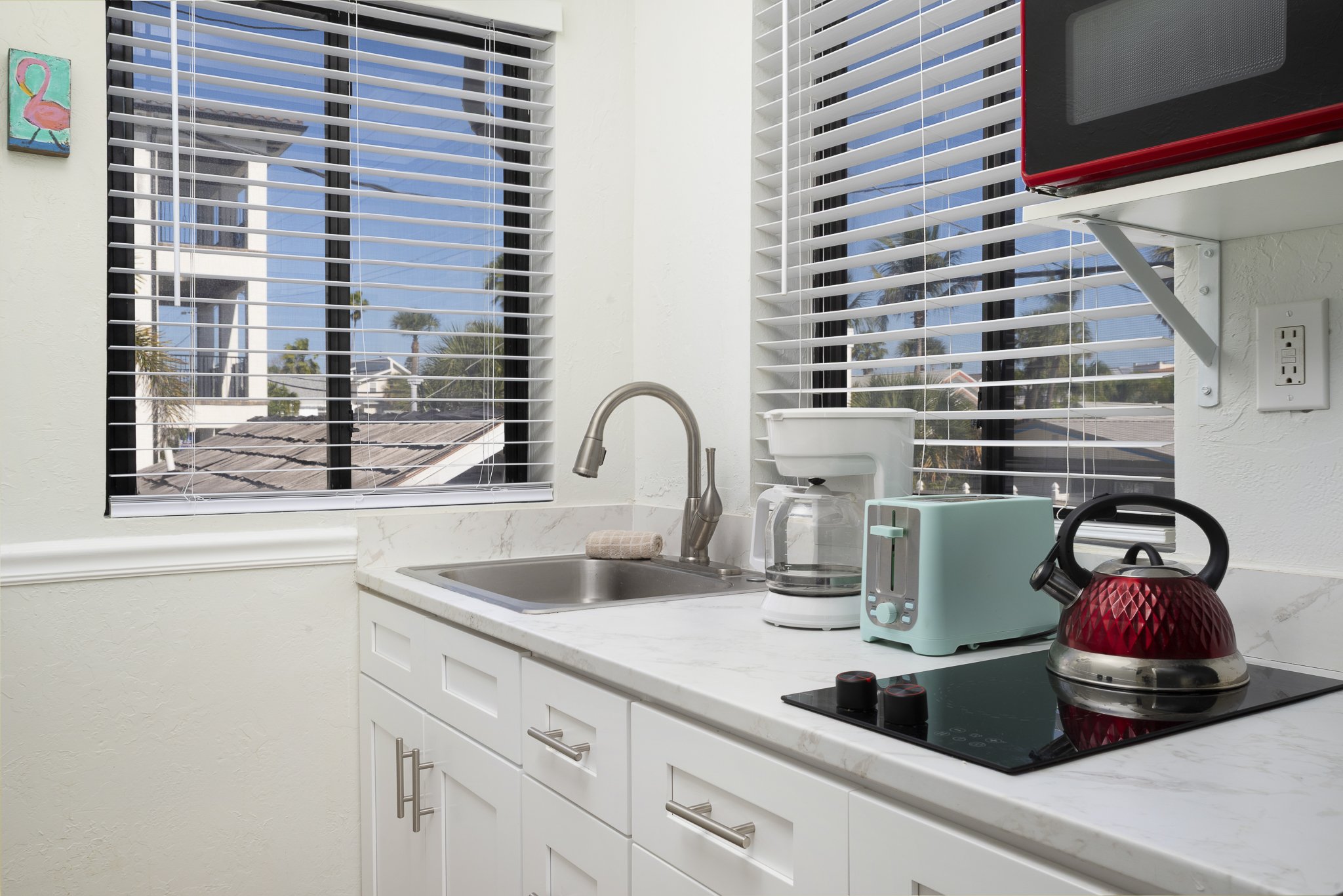 A kitchen countertop with a stainless steel sink, a coffee maker, a teal toaster, a red teapot on an electric stove, and white cabinets. Two large windows with blinds show buildings, palm trees, and a clear blue sky outside.