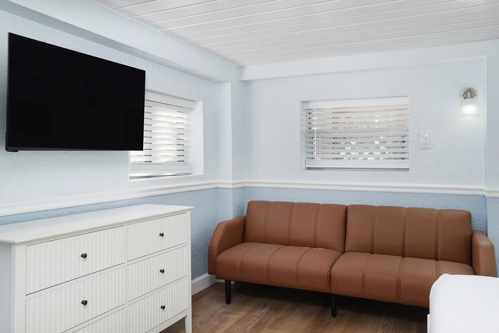 Living room with a wall-mounted flat-screen TV, two small windows with white blinds, a brown sofa, and a white dresser at Sabal Palms Inn.