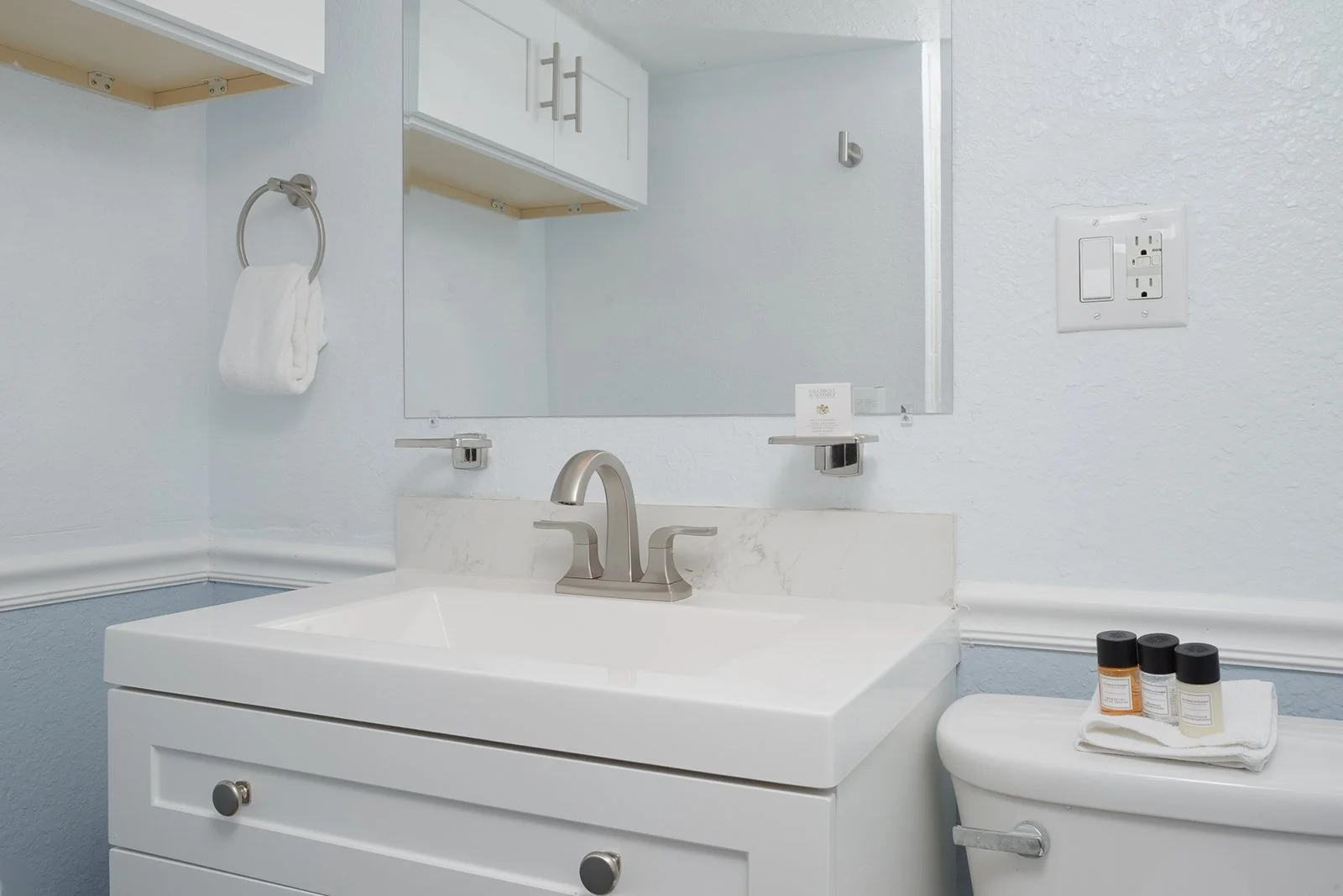 A clean bathroom sink with a white cabinet, a silver faucet, a mirror, a small shelf with toiletries, a towel ring with a white towel, and a toilet with a towel and small bottles on top.