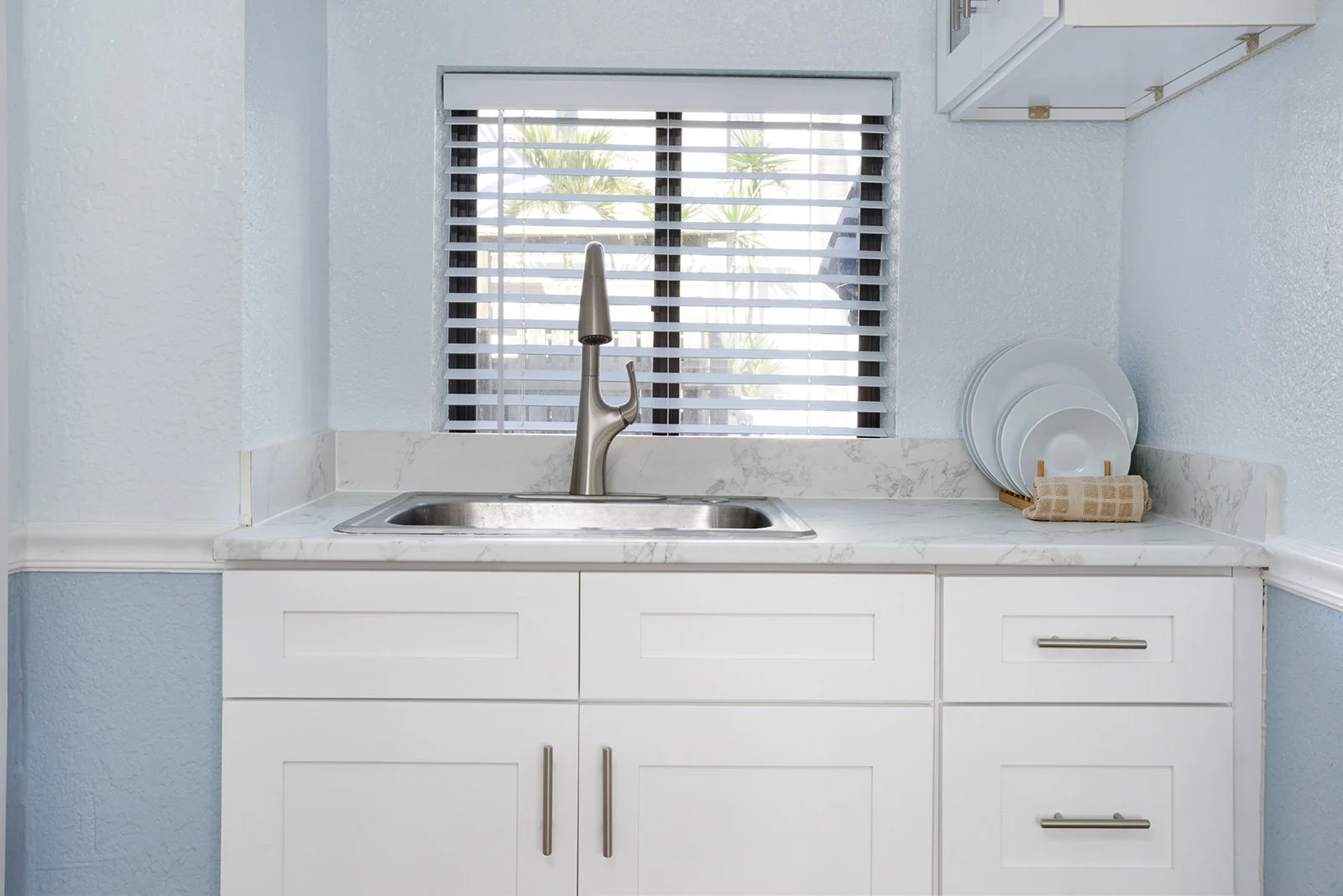 Kitchen sink with a window behind it, a faucet, white cabinets, and a set of white plates on a rack with a cloth, with palm trees visible through the window.