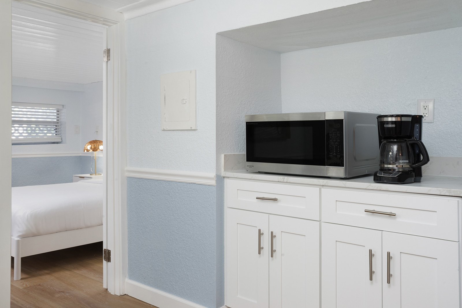 Kitchen area with microwave oven and coffee maker on white countertop, white cabinetry, and a doorway leading to a bedroom with a bed and a nightstand with a lamp at Sabal Palms Inn.