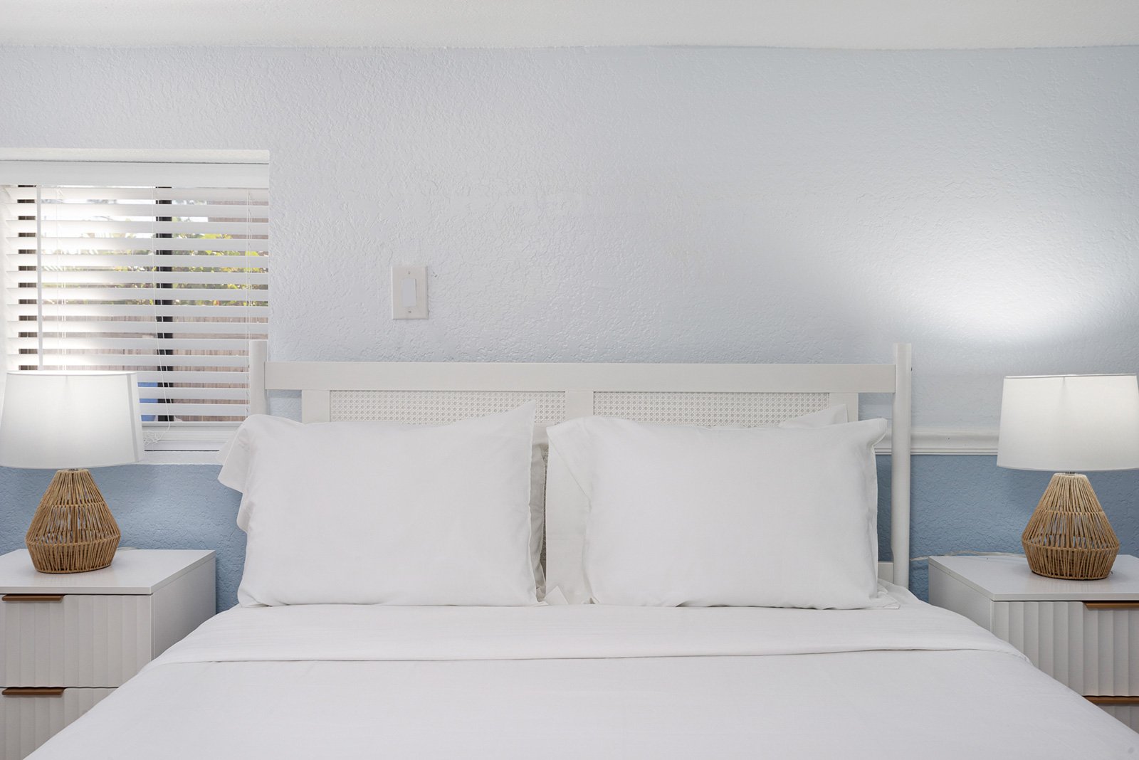 A neatly made bed with white sheets and pillows, two nightstands with wicker lamps, a window with horizontal blinds, and a textured white wall in a bedroom.