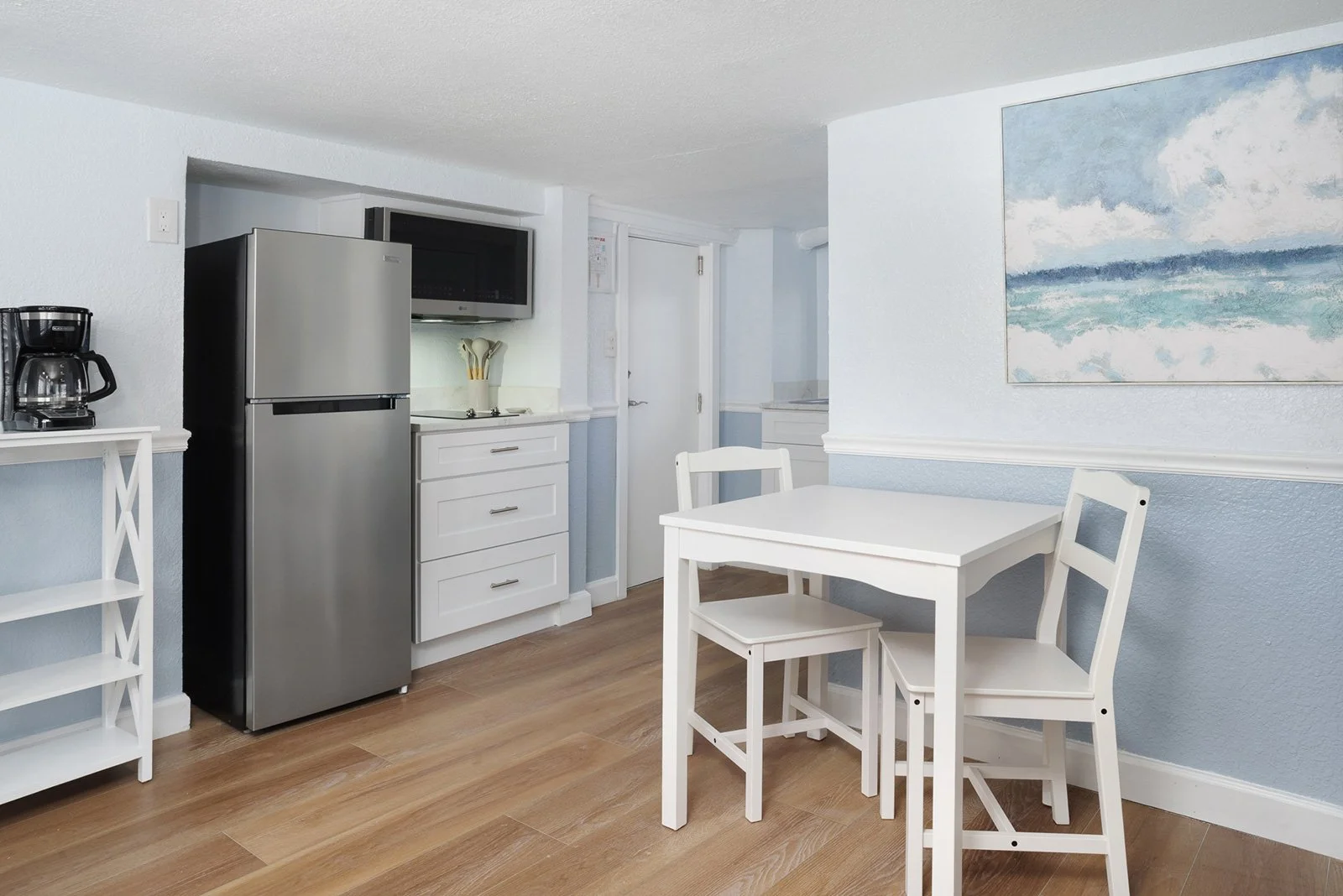 A compact kitchen area with a stainless steel refrigerator, a black microwave, a coffee maker, and white cabinetry. Adjacent to the kitchen is a small white dining table with two matching chairs, and a large abstract ocean painting on the wall.