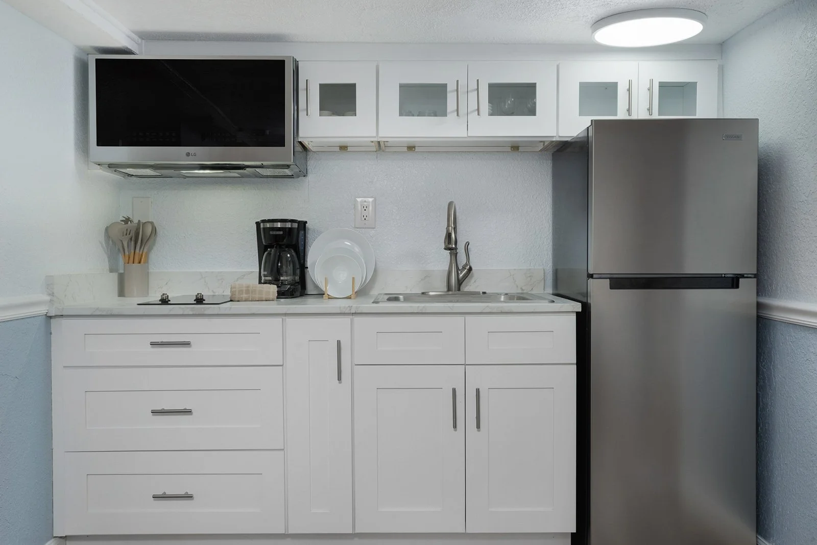 Small modern kitchen with white cabinets, a stainless steel refrigerator, a black coffee maker, two white plates, and a microwave mounted above.