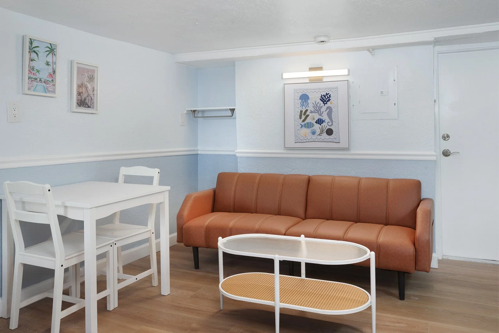 Living room with a brown sofa, white coffee table, and a small white dining table with chairs. The walls are painted light blue with framed pictures, and there is a door on the right side.