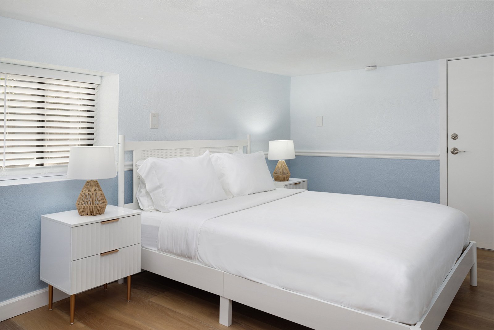 A minimalist white bed with white pillows and bedding, two wicker table lamps on white nightstands, a window with horizontal blinds, and a light blue wall in the background.
